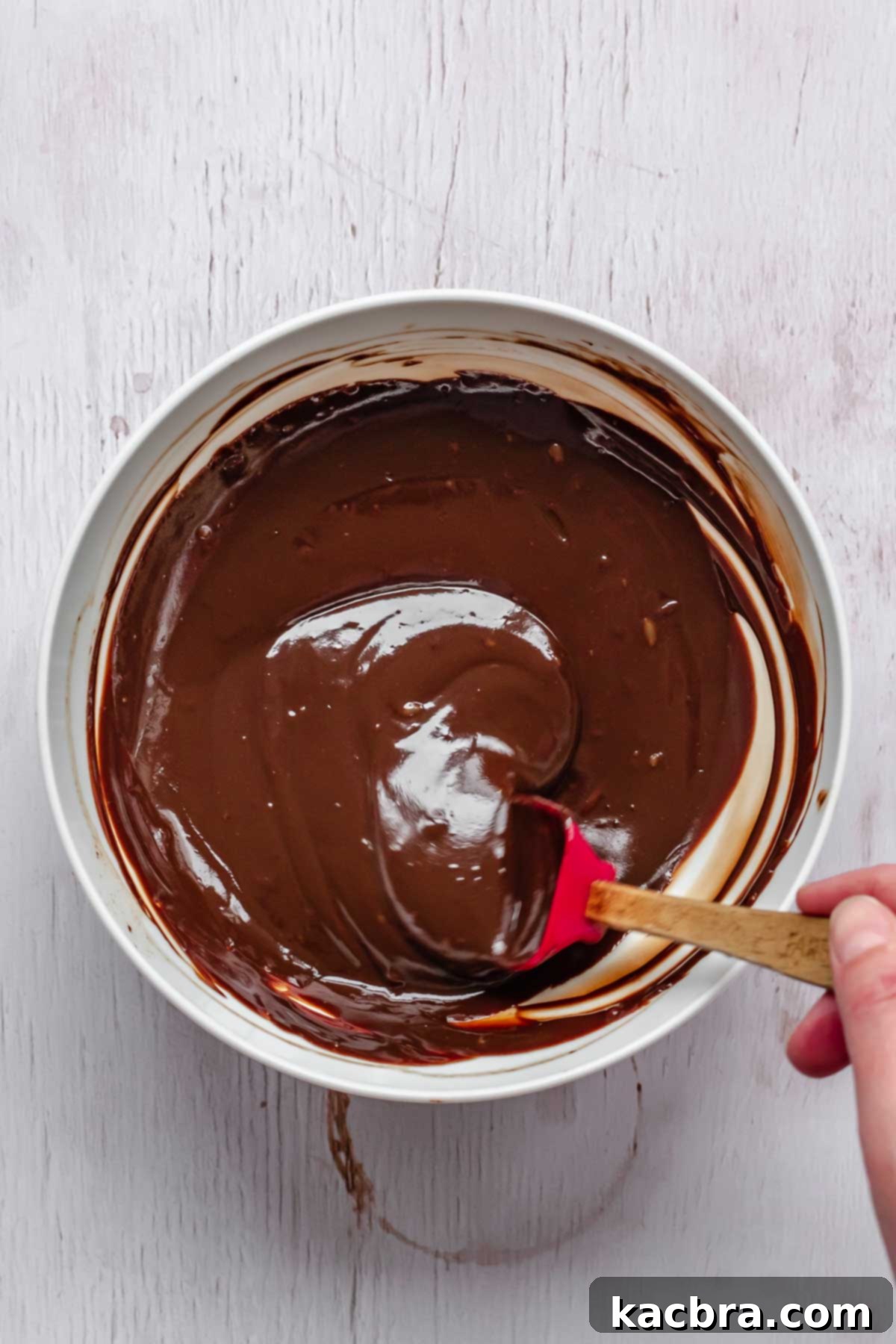 A mini spatula stirring rich, glossy chocolate ganache in a bowl, showing its smooth and pourable consistency after adding Baileys Irish Cream.