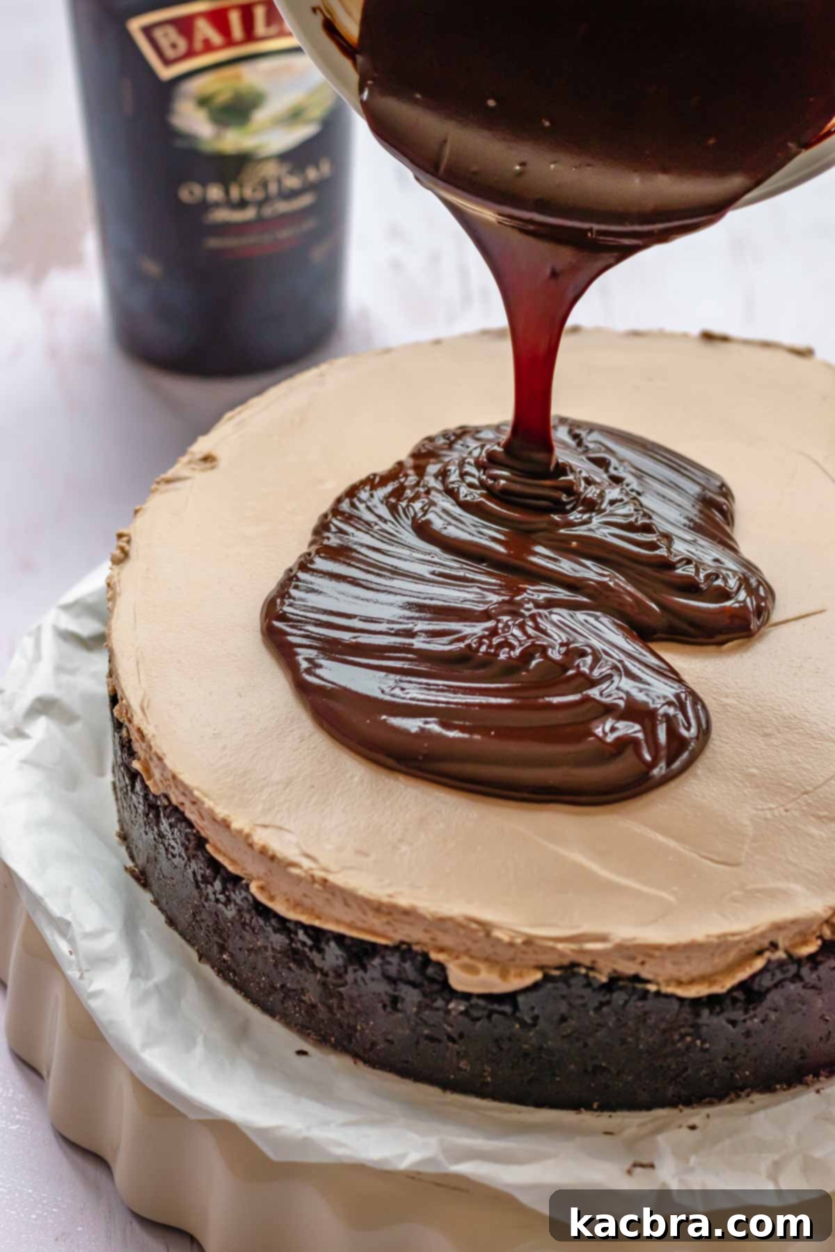 Warm, glossy chocolate ganache being poured over the top of a chilled no-bake cheesecake, slowly dripping down the sides for a beautiful effect.