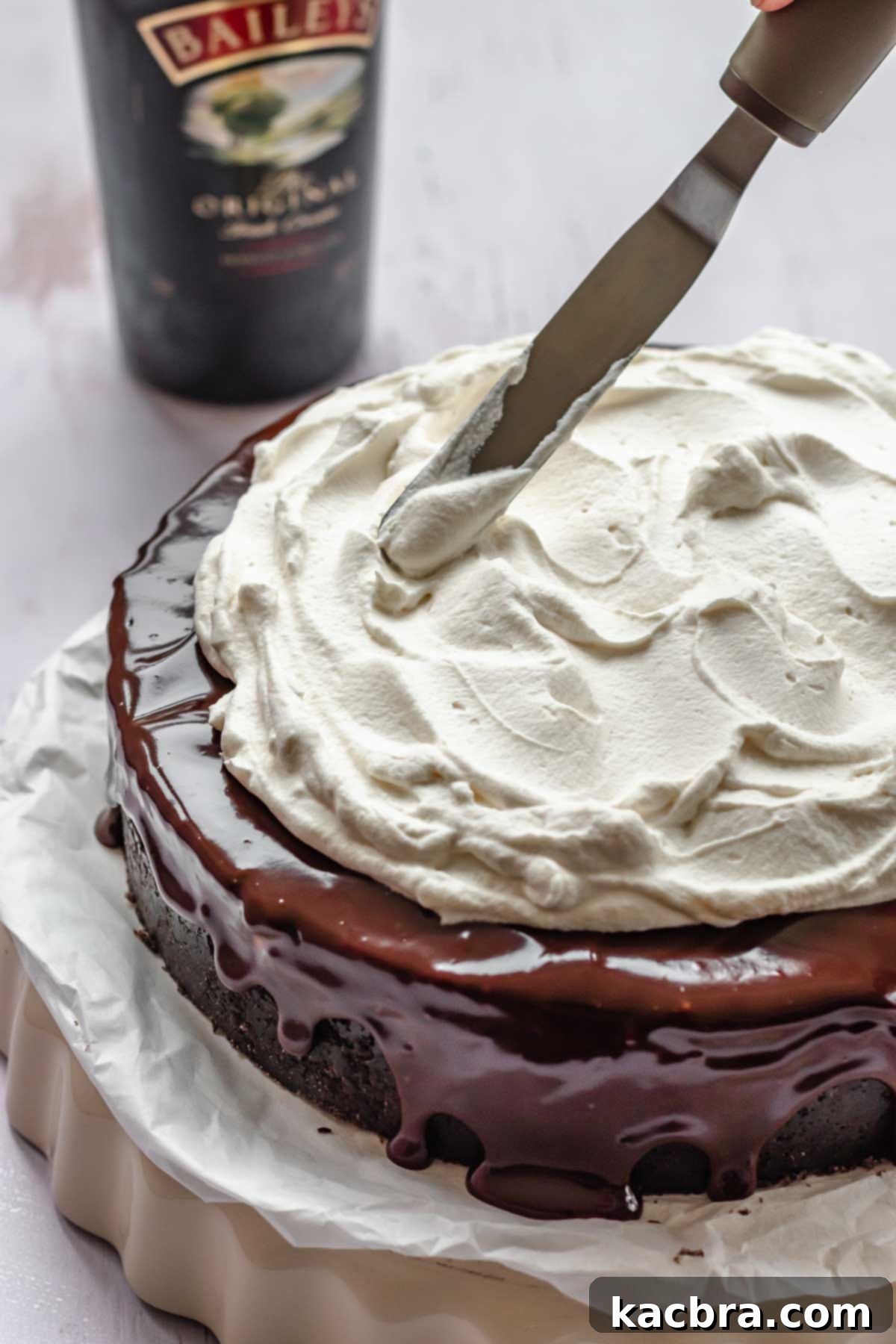 An offset spatula artfully adding fluffy Baileys whipped cream in decorative swoops on top of the chocolate-ganache-covered cheesecake, ready for garnish.