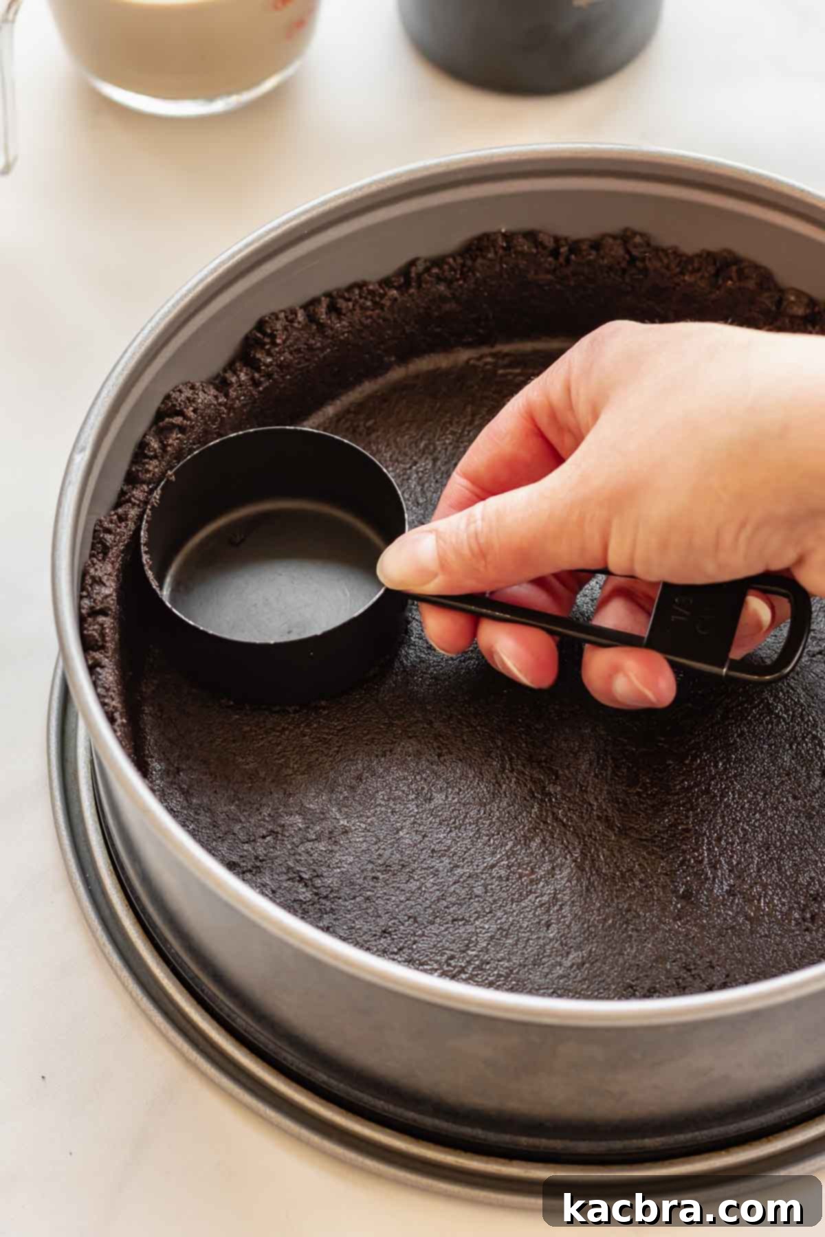 A hand pressing the moist Oreo crumb mixture firmly into the bottom and halfway up the sides of a springform pan using a dry measuring cup.