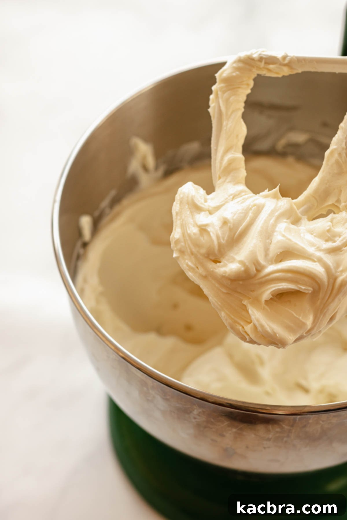 Softened cream cheese and granulated sugar being creamed together with a paddle attachment in a stand mixer bowl, showing a light and fluffy texture.