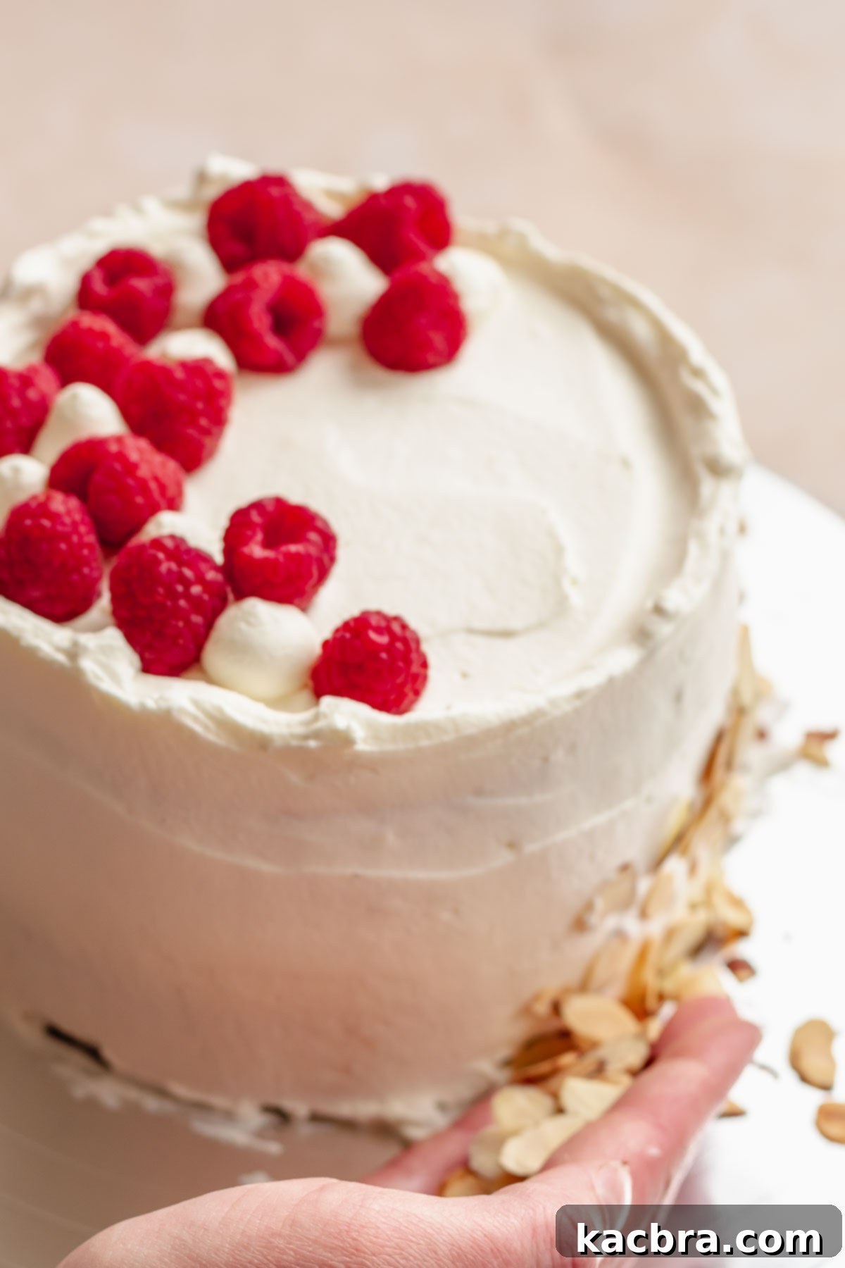 Crimson Kissed Almond Delight 18 A hand pressing sliced almonds onto the base of the cake, with fresh raspberries arranged on top.