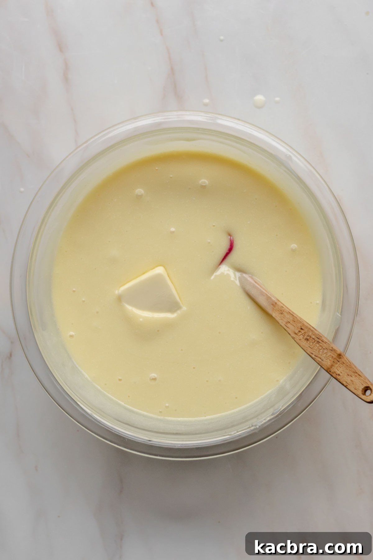 A pat of room temperature butter being added to the warm white chocolate ganache, with a spatula resting in the bowl.