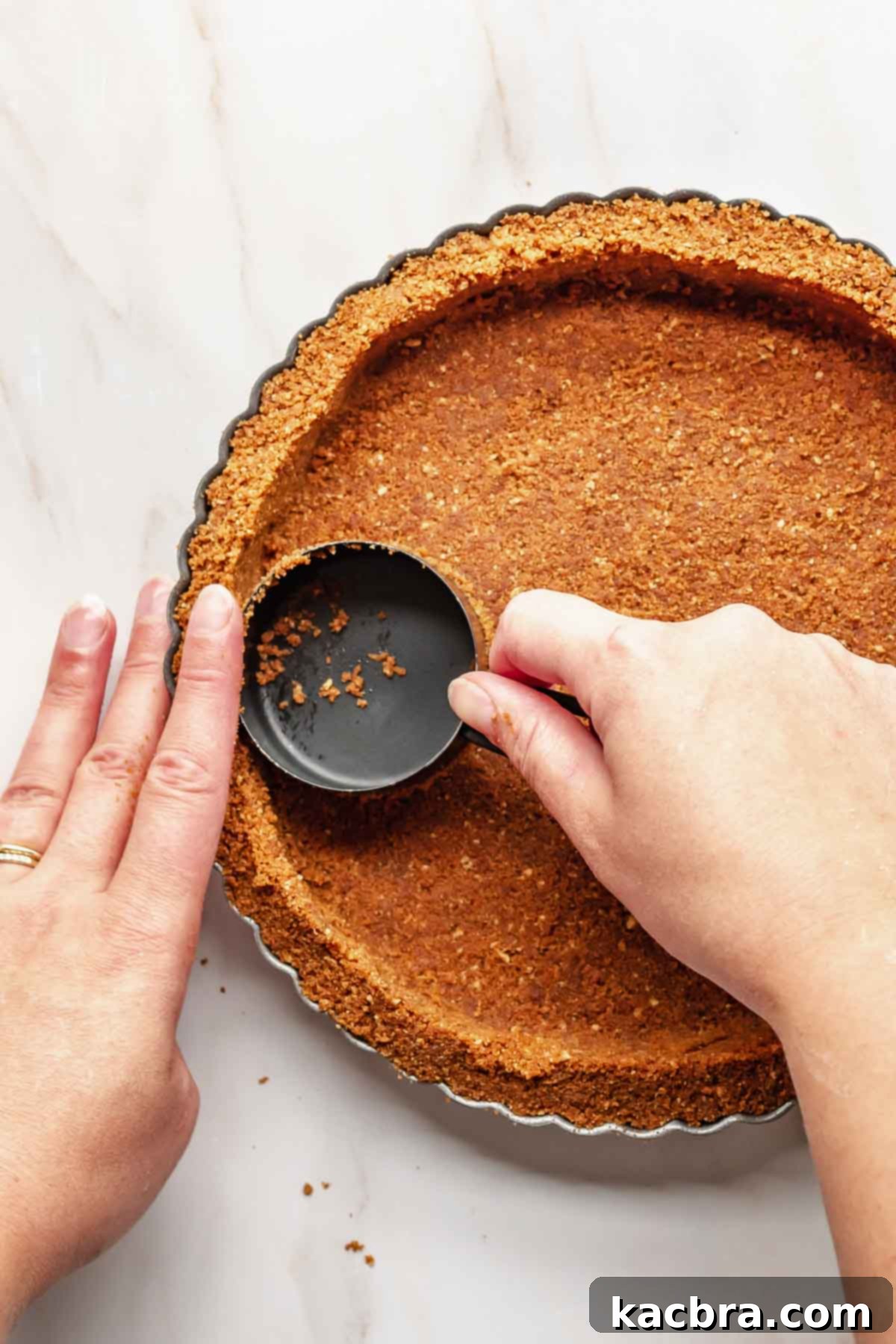 Hands pressing the Biscoff cookie crumb mixture firmly into a tart pan with a removable bottom, using a measuring cup.