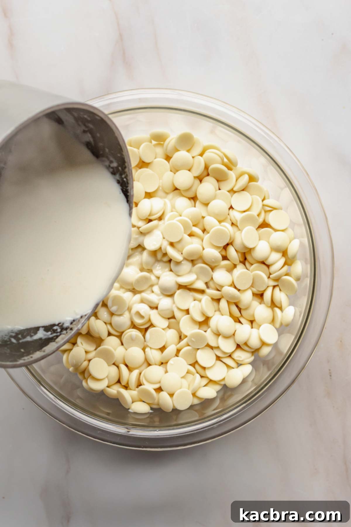 Steaming heavy cream being poured over finely chopped white chocolate in a bowl to create the ganache base.