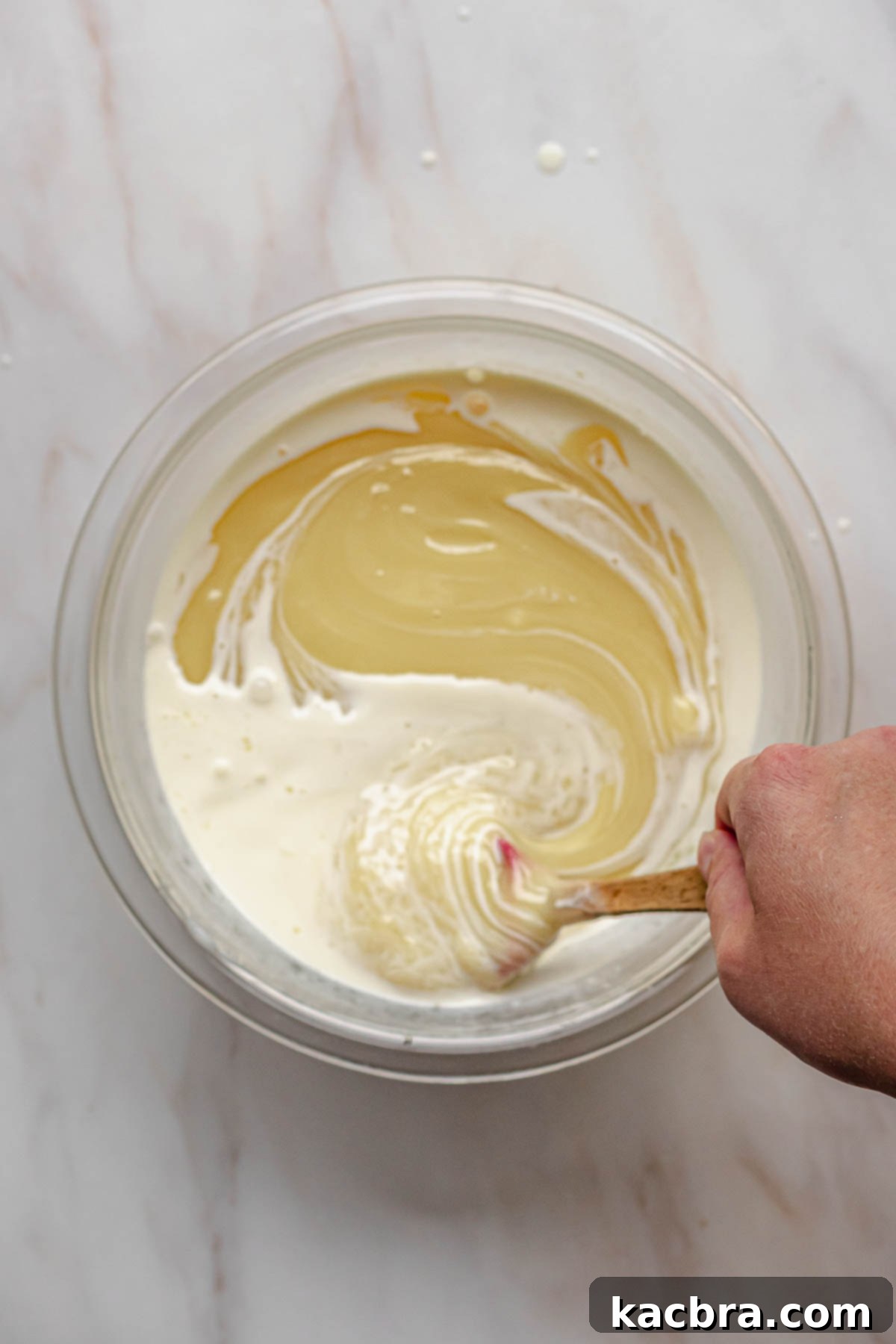 A hand gently stirring melted white chocolate and heavy cream with a rubber spatula until a smooth ganache forms.