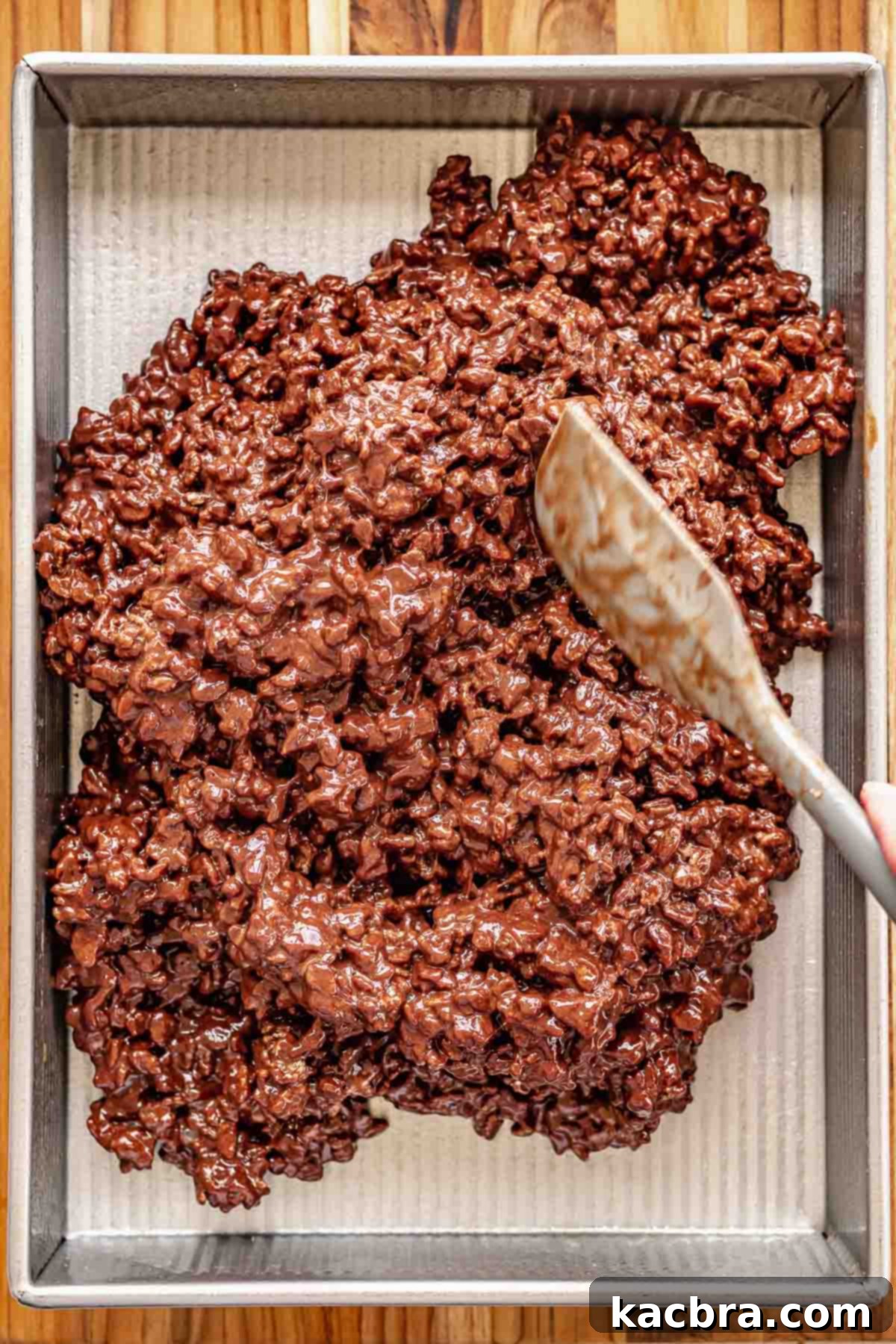 A spatula spreading the chocolate Rice Krispie mixture evenly into a parchment-lined pan.