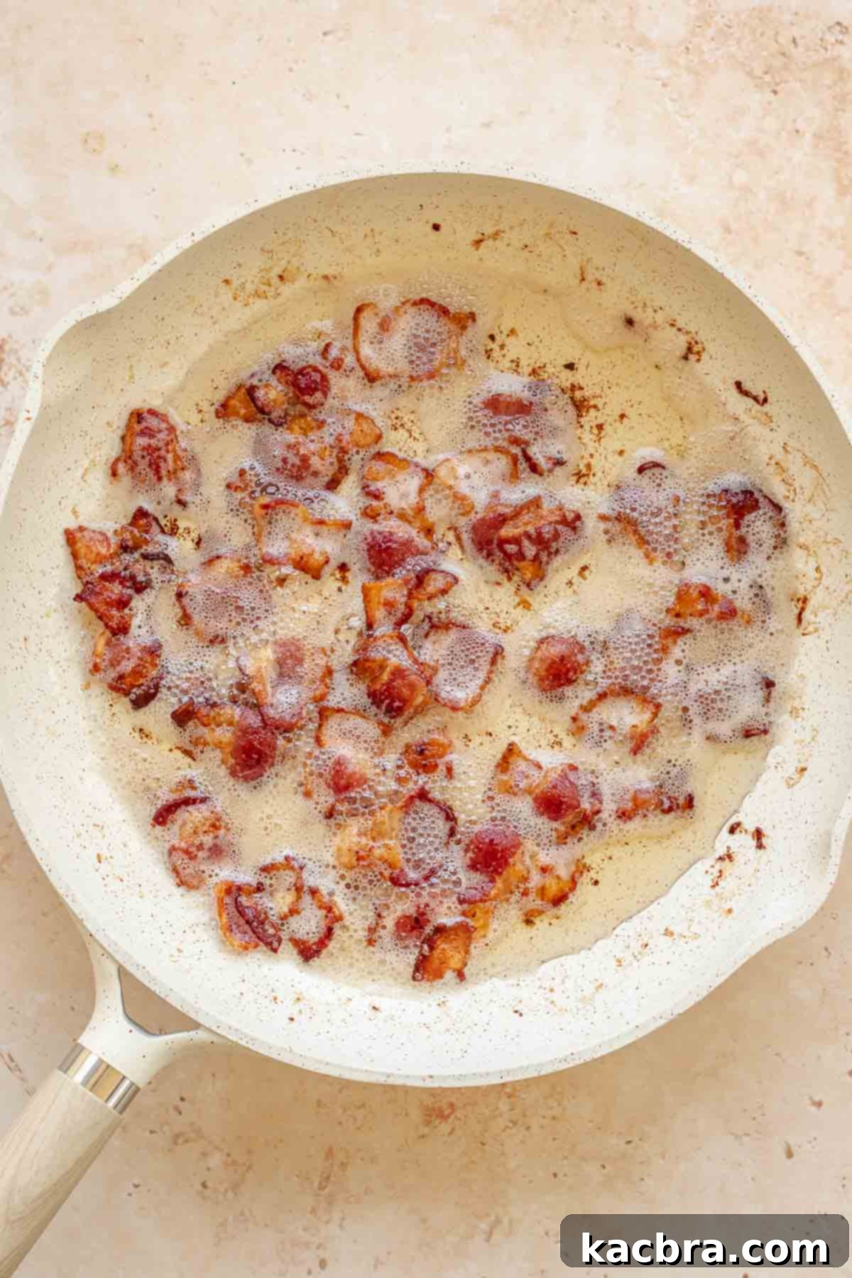 Bacon pieces cooking in a pan, rendering fat and becoming crispy.