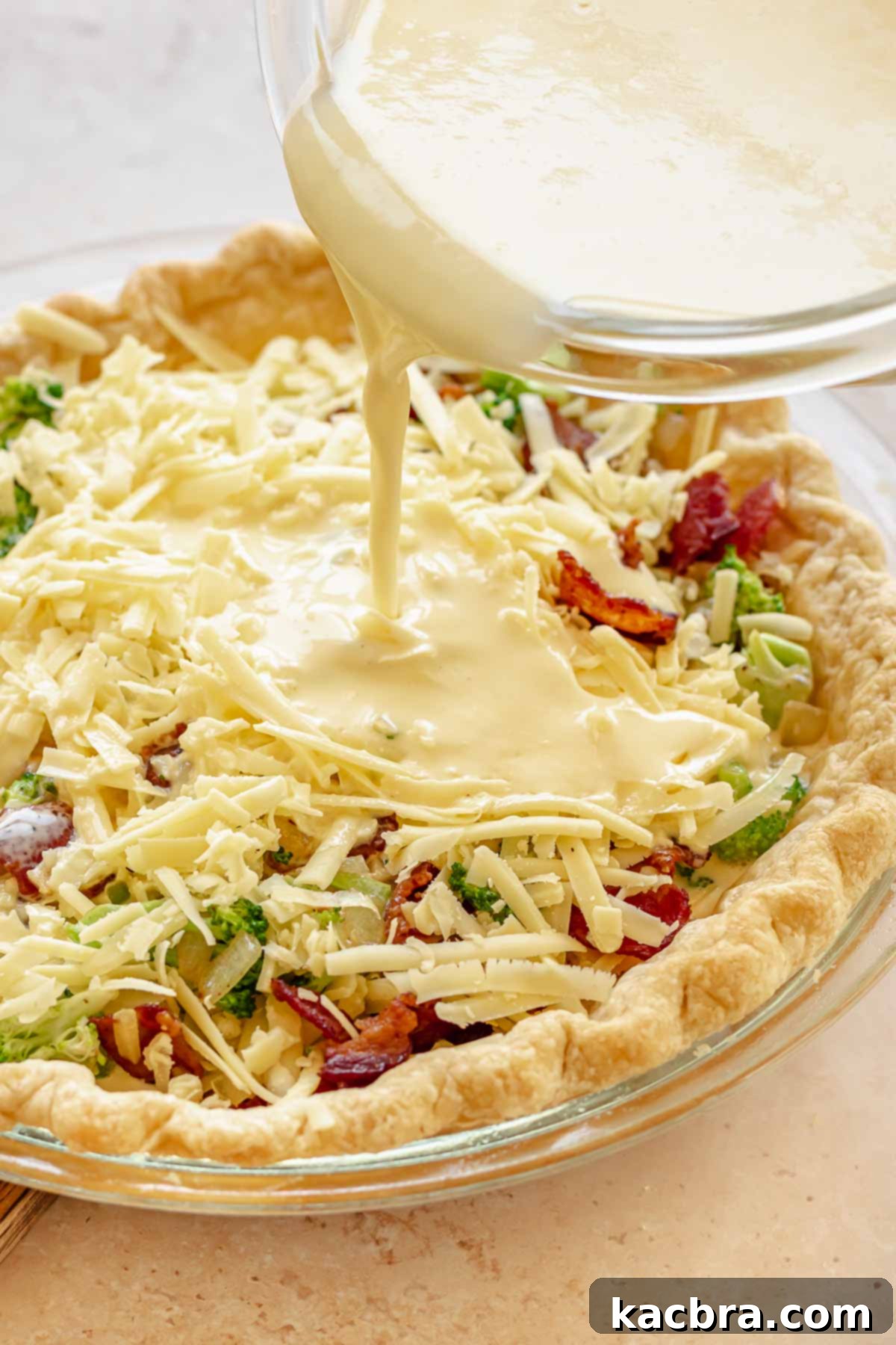 Egg custard being carefully poured over the bacon, broccoli, and cheese filling in the pie crust.