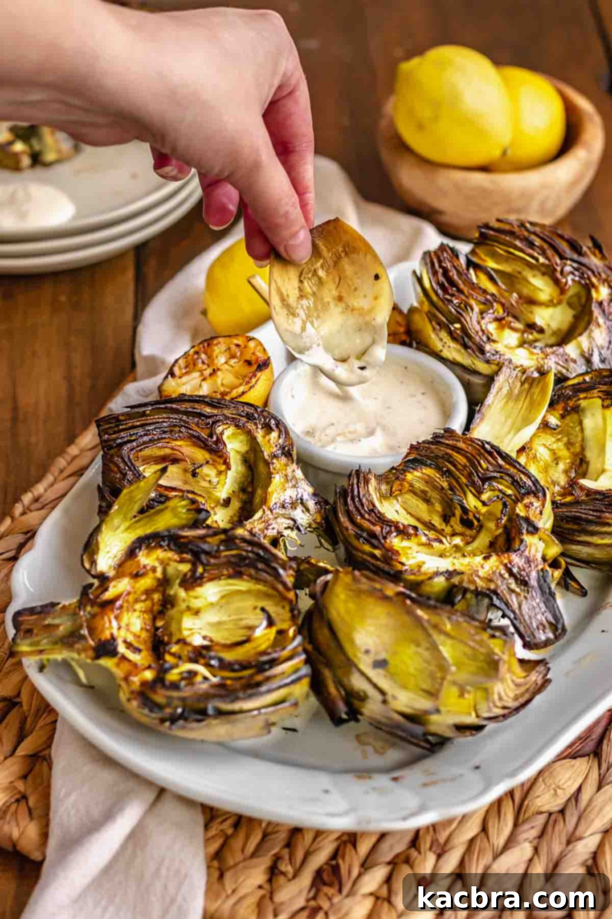 A hand dips an artichoke leaf into aioli. Other artichokes sit on a platter.