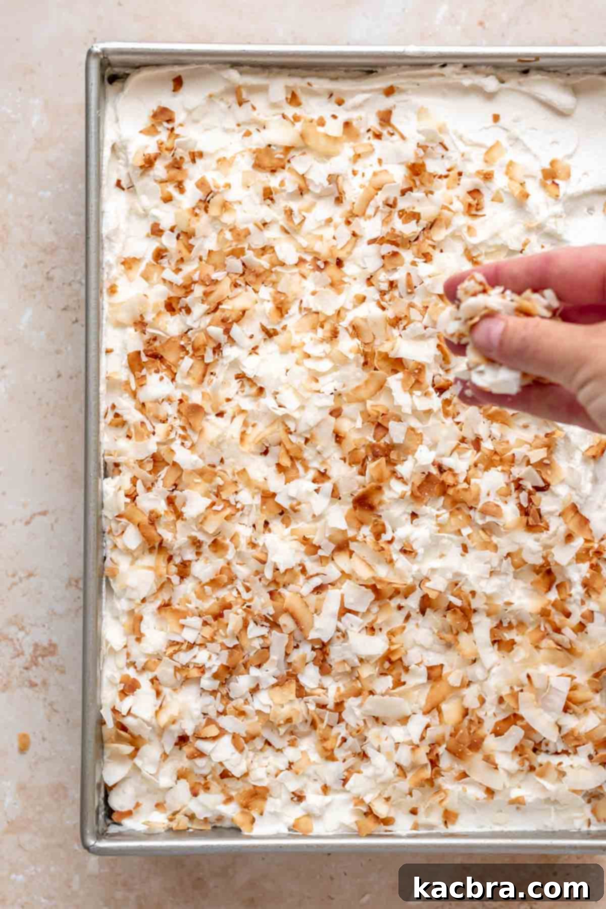 A hand sprinkles toasted coconut flakes over the whipped cream on the cake.