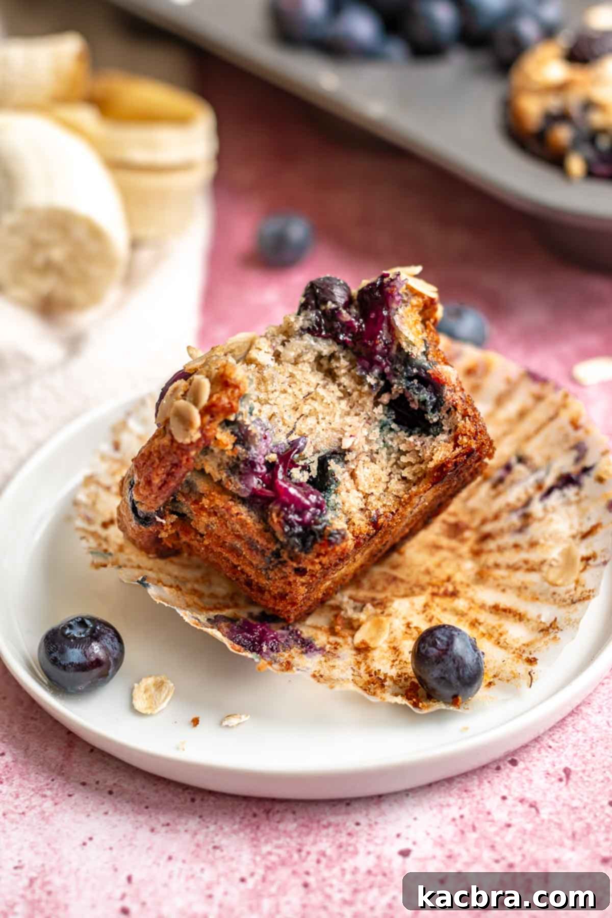 Sweet and Healthy Banana Blueberry Oatmeal Muffins 2 A golden-brown blueberry muffin on a white plate, with a bite taken out revealing the soft, fruity interior.