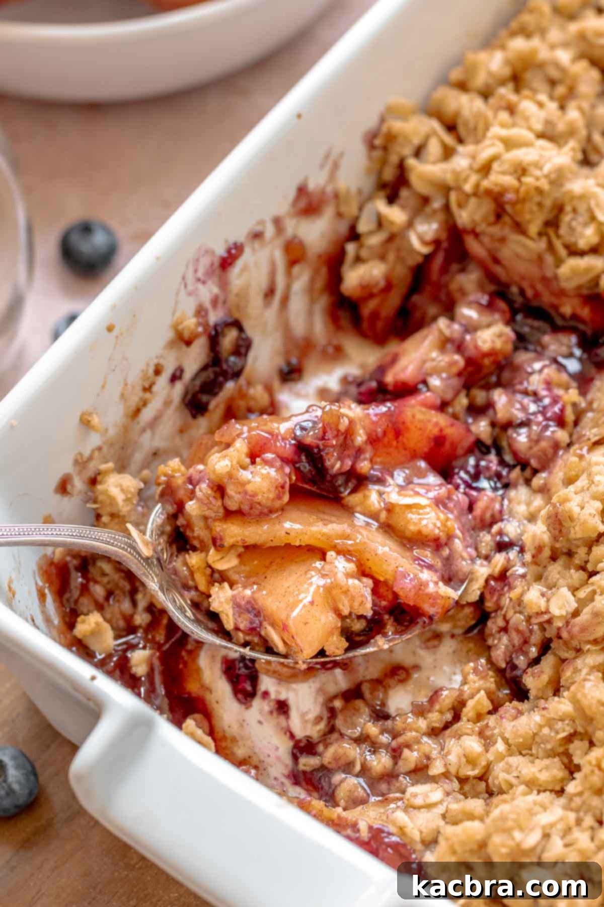 A scoop of apple blueberry crisp on a spoon sitting in a casserole dish, garnished with vanilla ice cream.