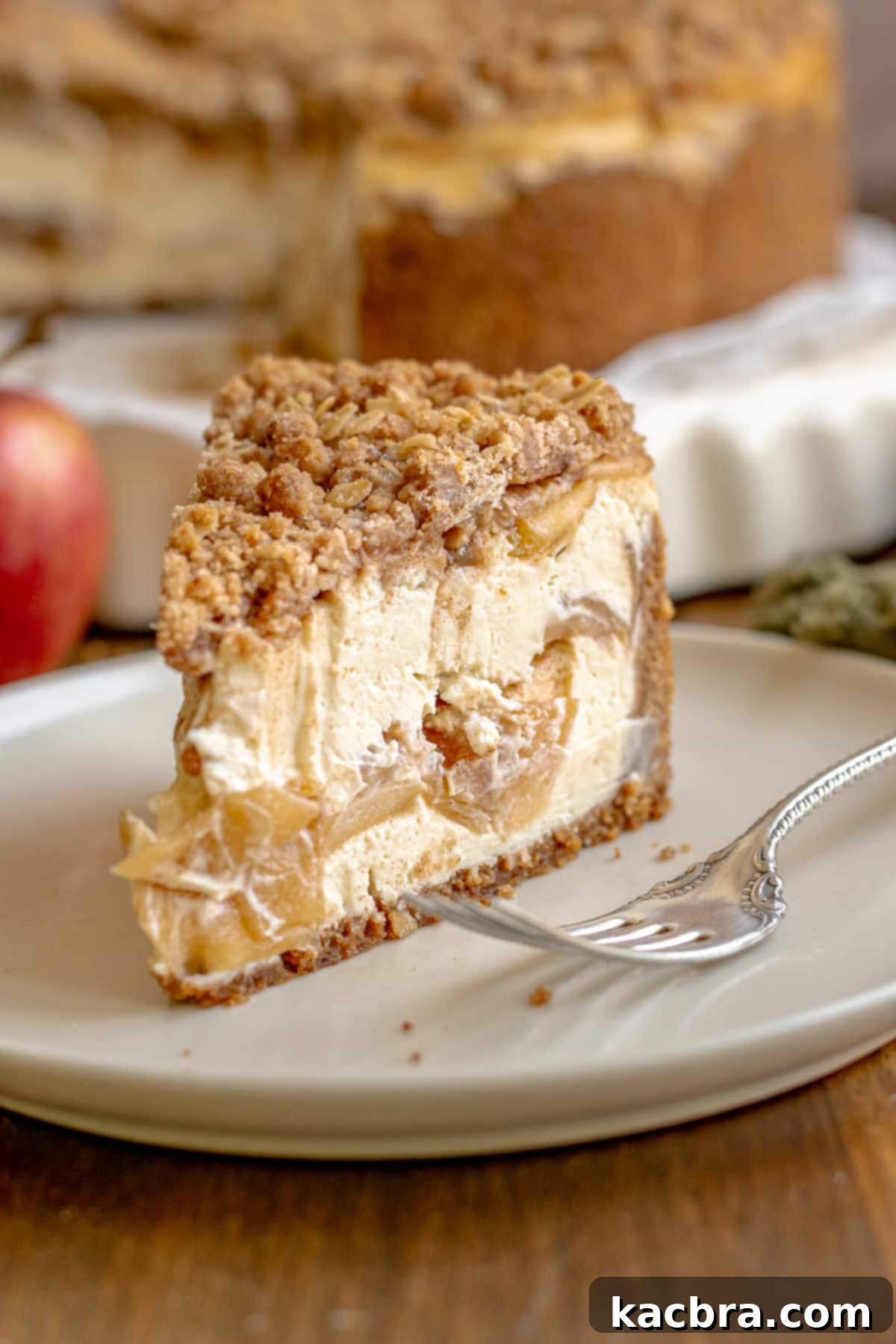 A slice of apple crisp cheesecake on a plate with a fork next to it.