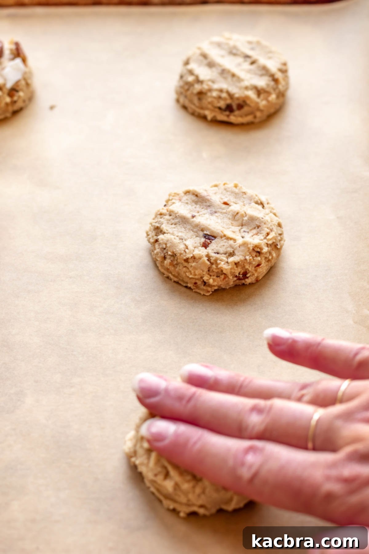 A hand gently pressing down on a scooped cookie dough ball on a baking sheet, slightly flattening it before baking.