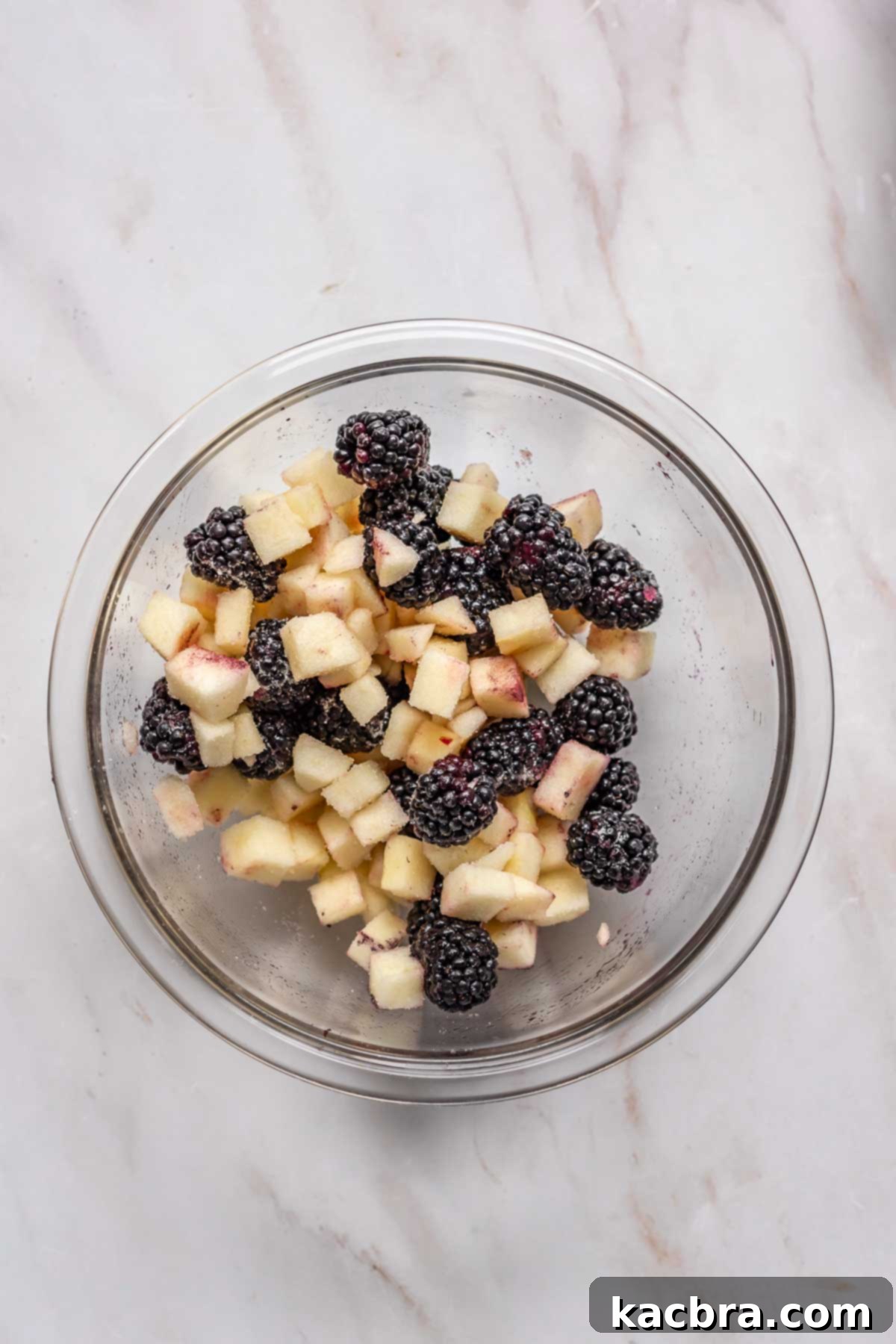 Diced apples and fresh blackberries being tossed with sugar in a mixing bowl.
