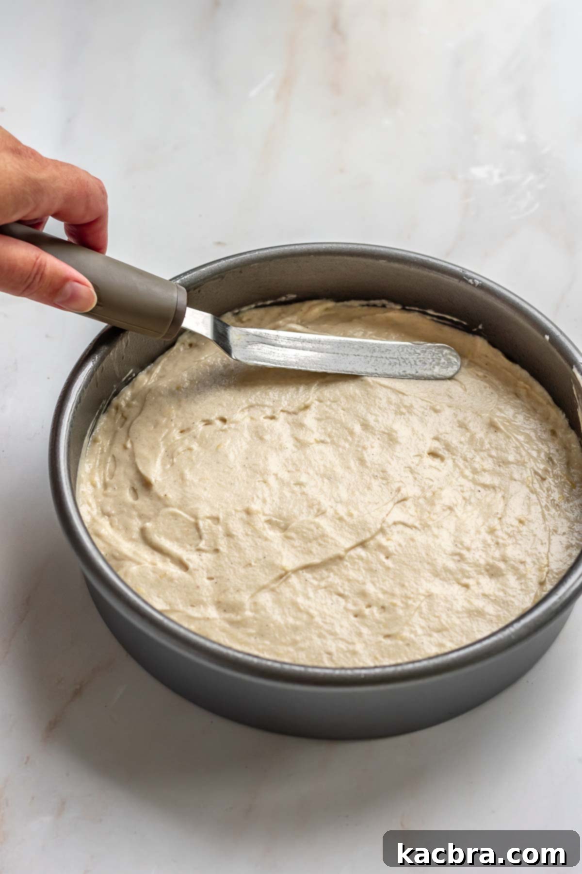 A hand spreading cake batter evenly into a prepared cake pan with an offset spatula.