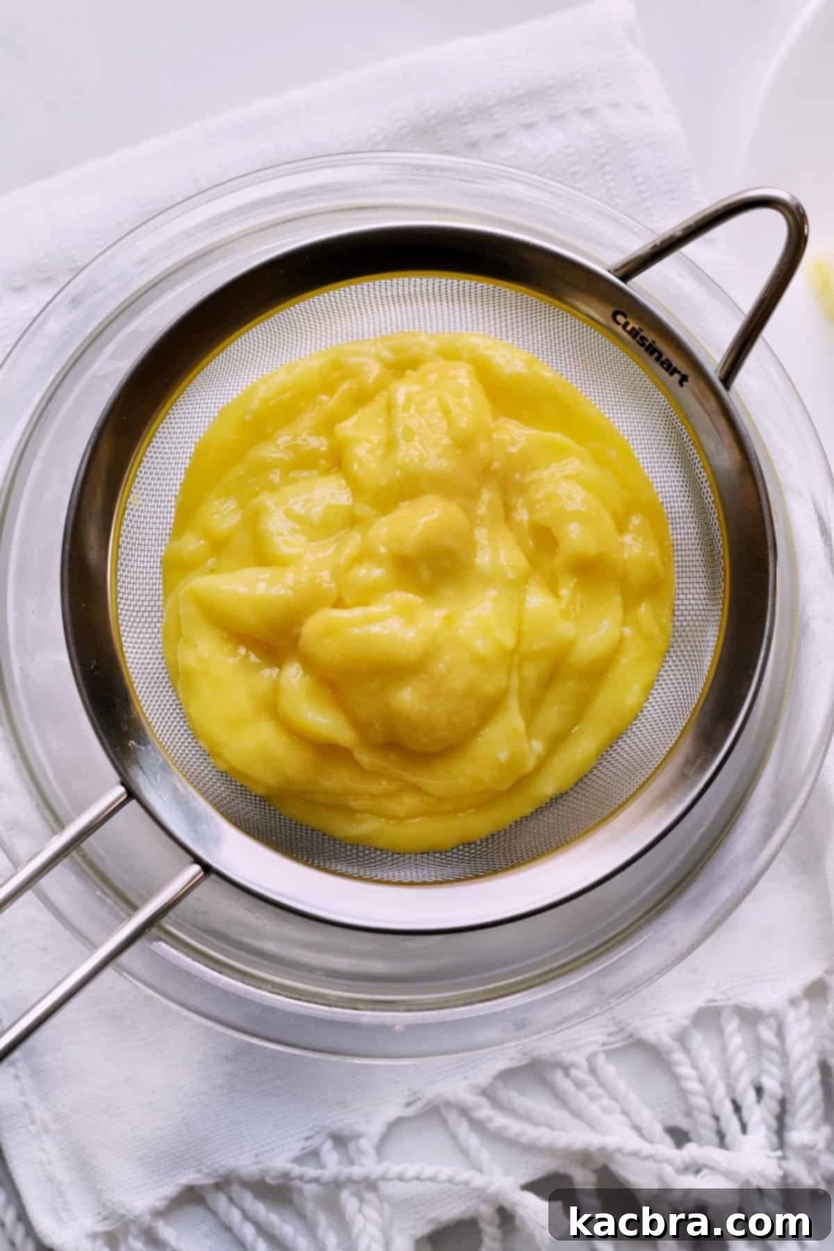 Hot, freshly cooked lemon curd being carefully poured into a fine mesh sieve placed over a bowl, ready for straining.
