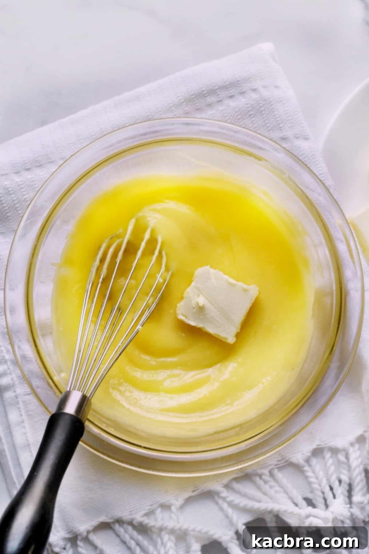 Room temperature butter pieces being whisked into a bowl of warm, strained lemon curd, creating a smooth and glossy finish.