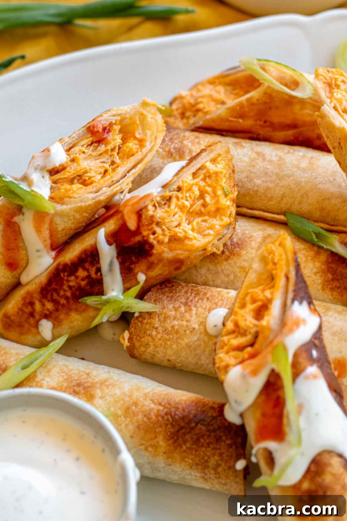 Platter of golden-brown Buffalo chicken taquitos, some cut open to reveal creamy filling, drizzled with ranch dressing.