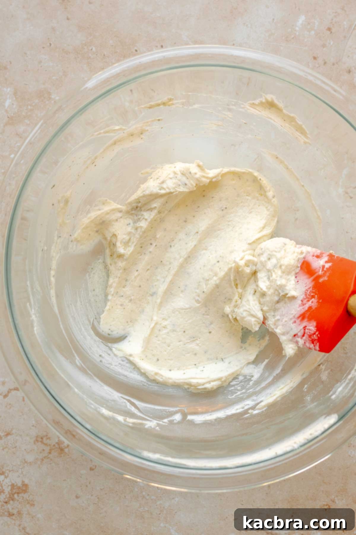 Softened cream cheese being mashed in a bowl with a rubber spatula.