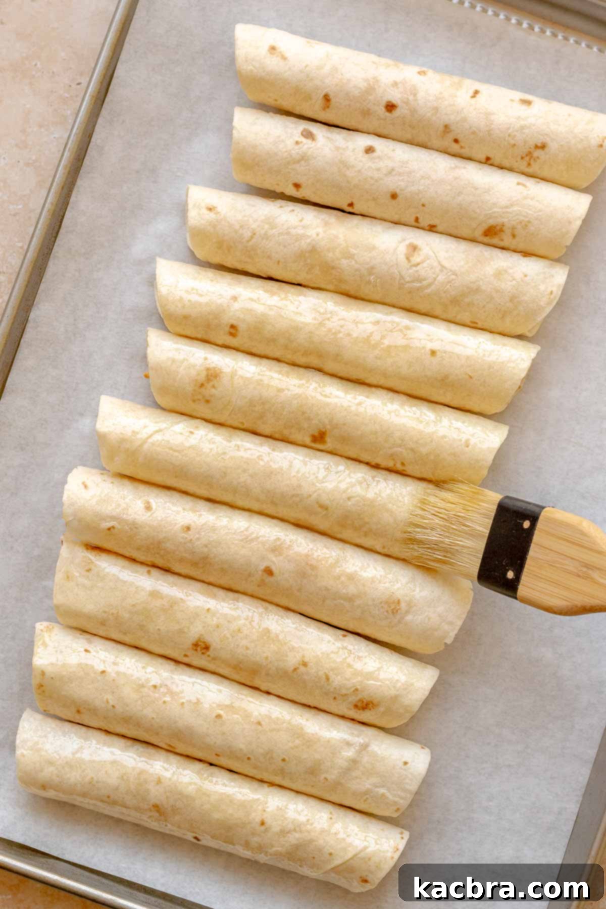 A pastry brush applying olive oil to rolled buffalo chicken taquitos on a baking sheet.