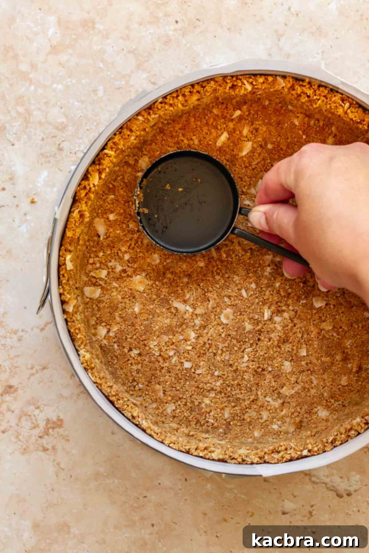 A hand uses a measuring cup to press graham cracker crust into a pan.