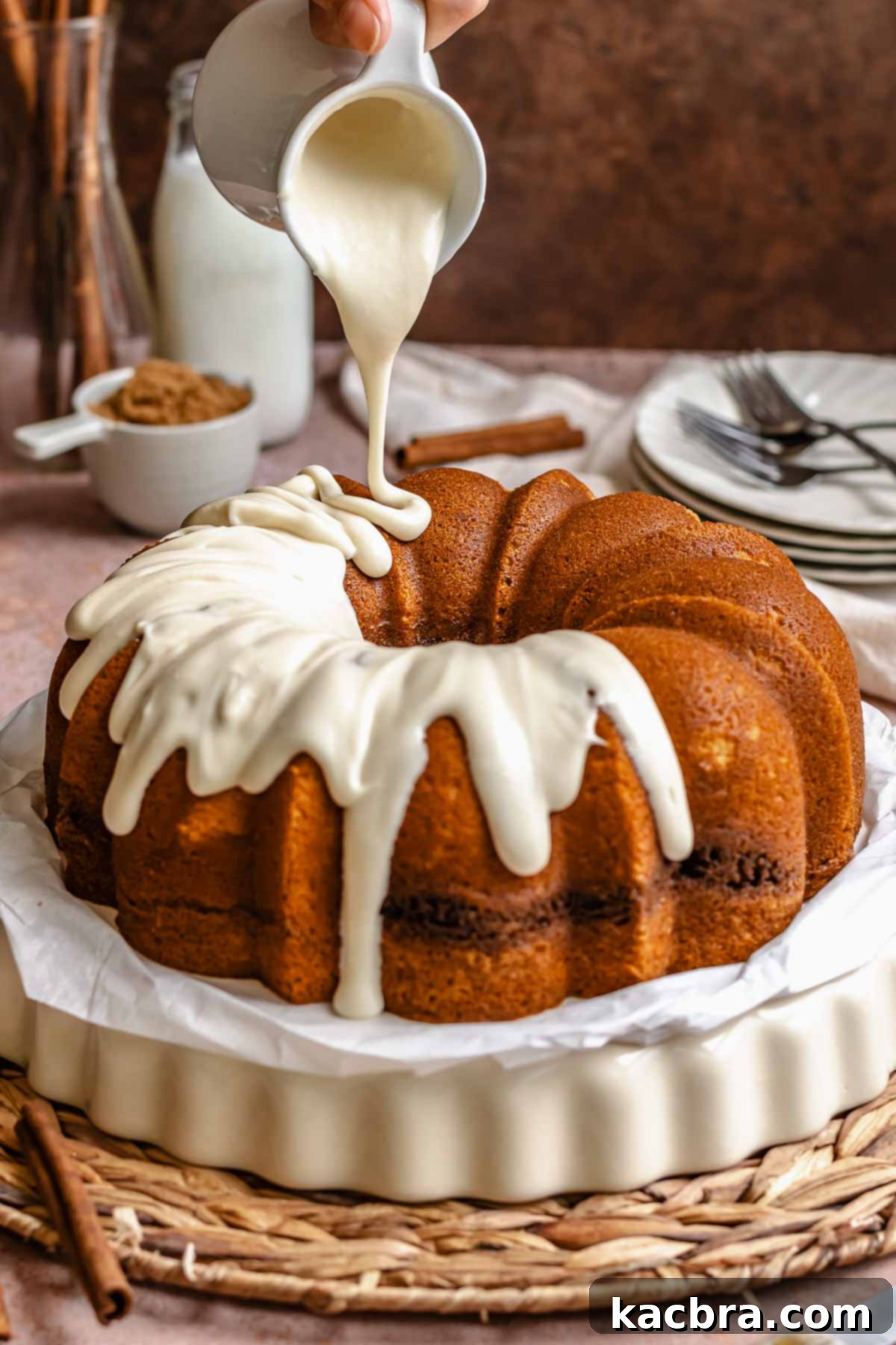 A hand pours cream cheese frosting over a cinnamon bundt cake on a platter.