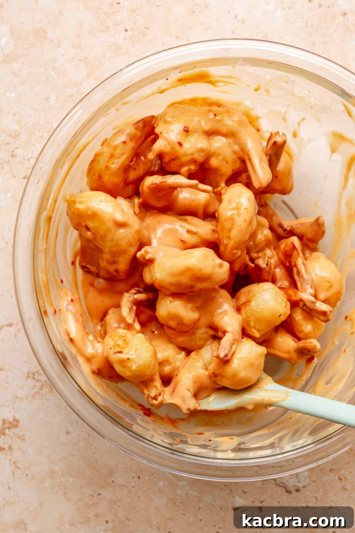 A spatula tosses fried shrimp with boom boom sauce in a bowl.