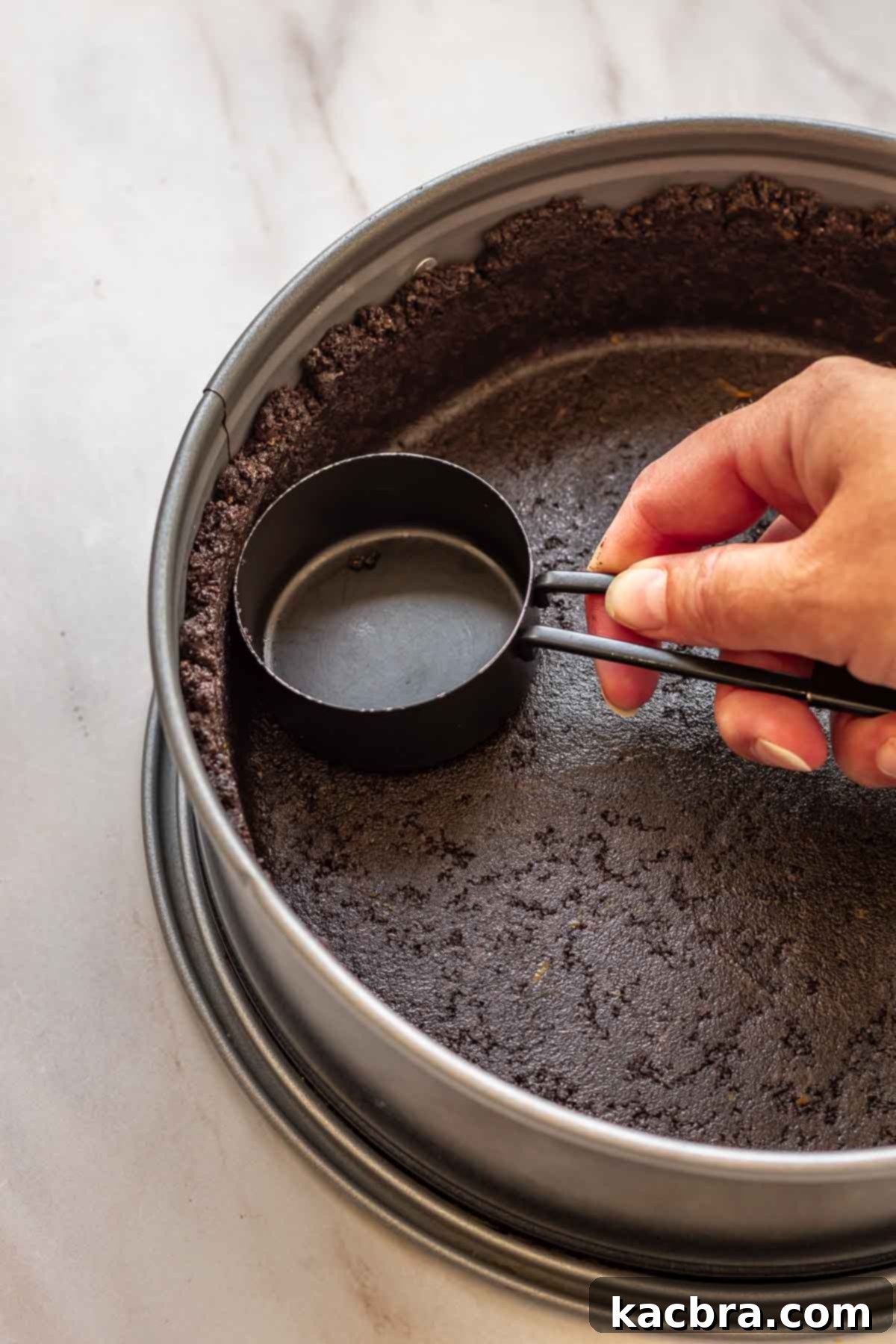 No-Bake Chocolate Orange Velvet Cheesecake 7 A hand uses a measuring cup to press oreo crumbs into a springform pan.