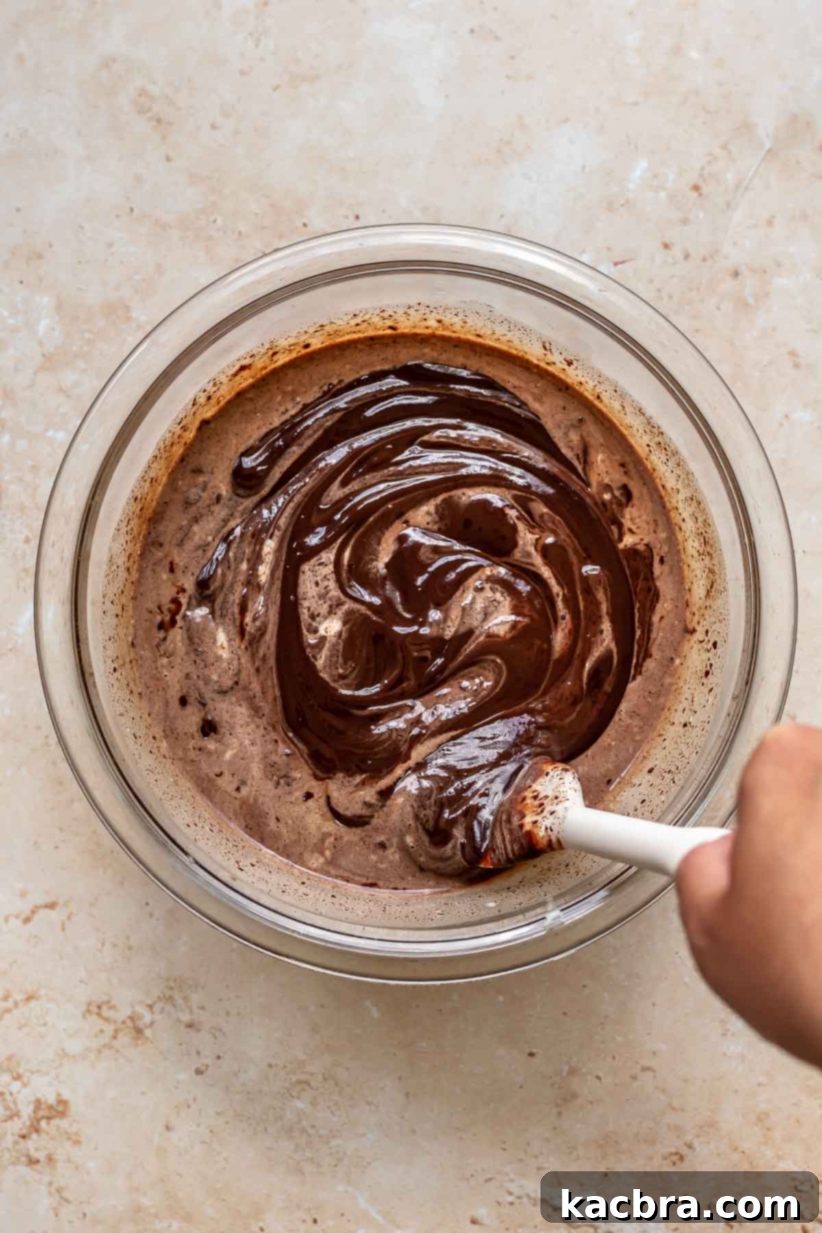 A rubber spatula mixes together hot cream and chopped dark chocolate to form a smooth ganache in a bowl.