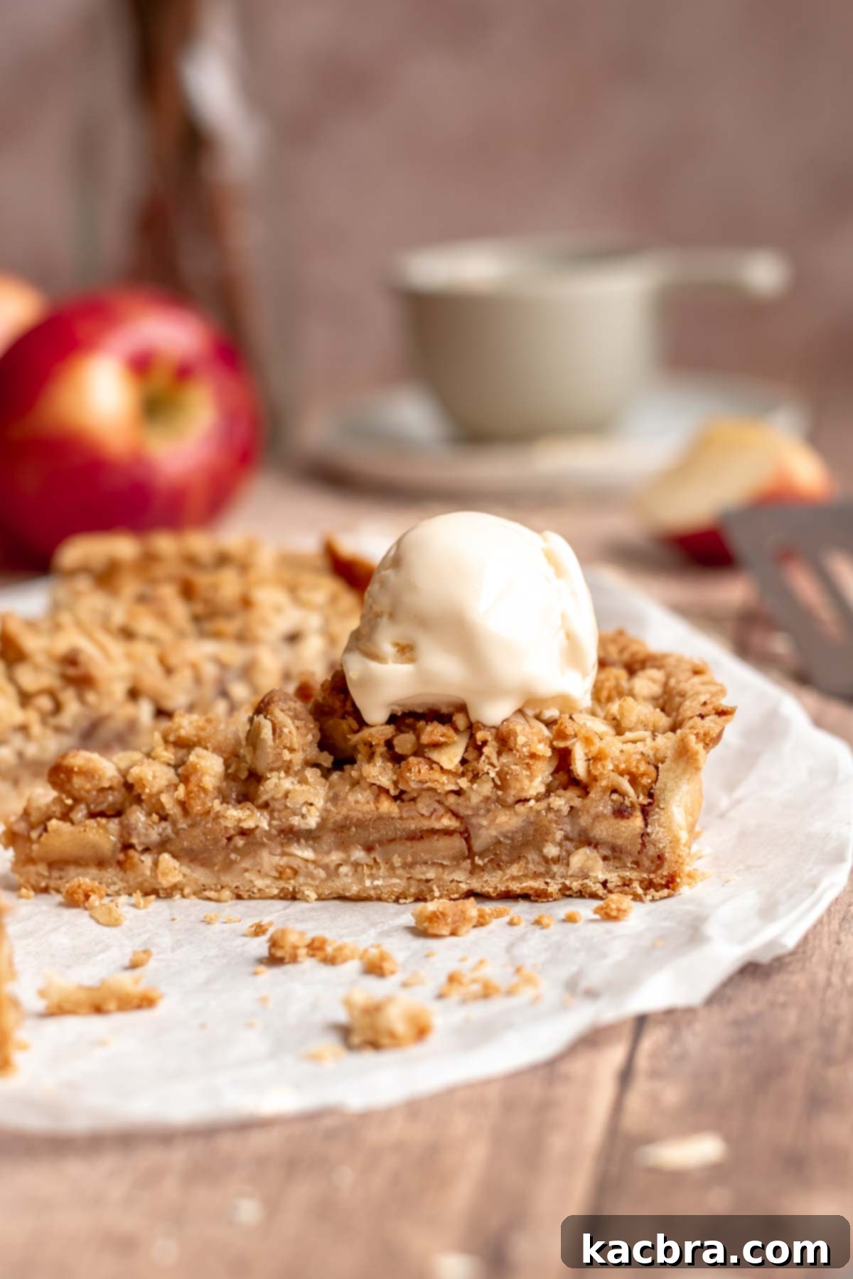 Cinnamon Apple Crumble Tart 2 A beautifully baked apple crumble tart on parchment paper, with one slice served and topped with a scoop of vanilla ice cream, showcasing its golden crumble and tender apple filling.