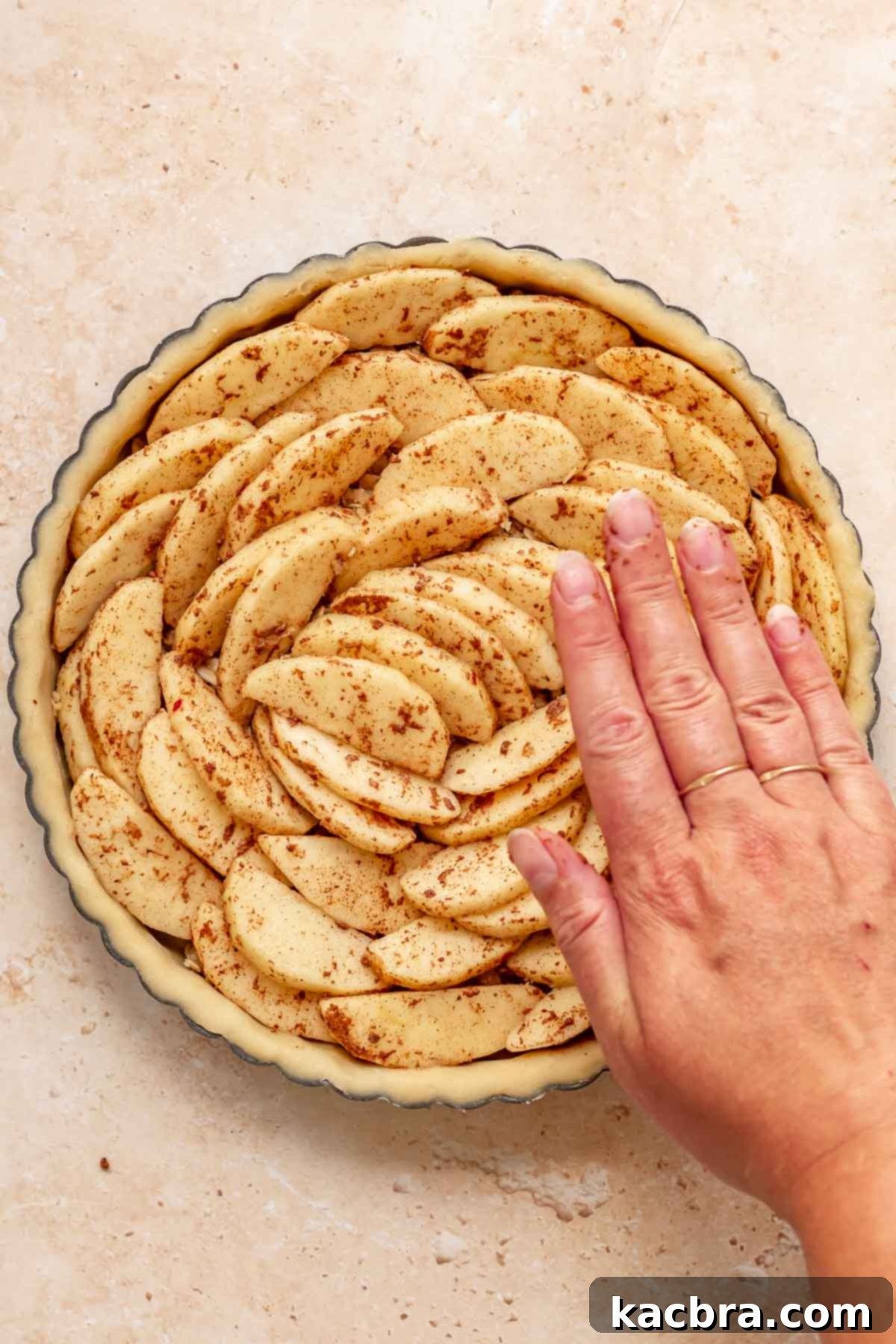 Cinnamon Apple Crumble Tart 11 A hand gently presses down on beautifully arranged apple slices in a concentric spiral pattern within a tart pan, maximizing the apple filling.