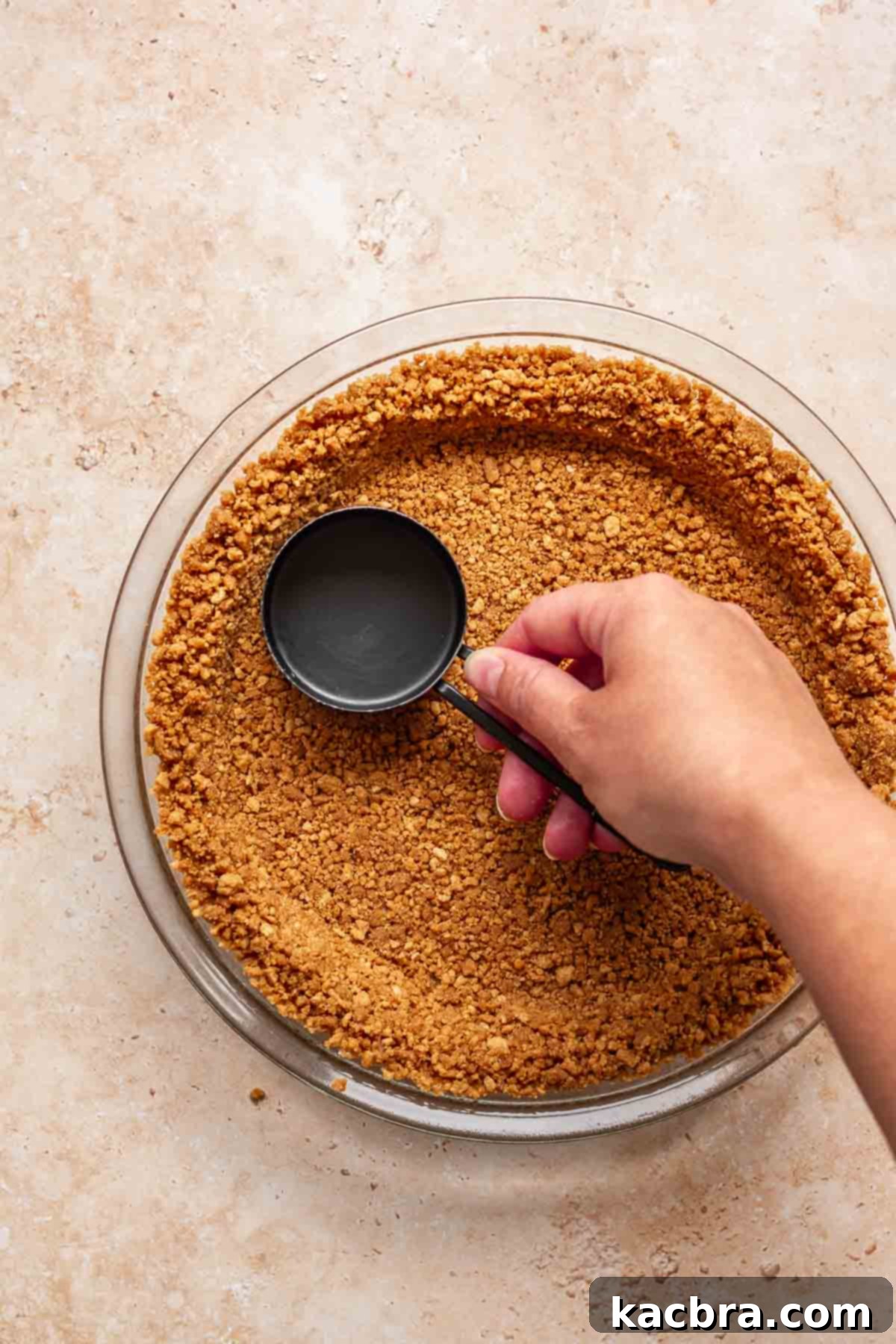 Creamy Pumpkin Cloud 5 A hand pressing the gingersnap crumb mixture firmly into a pie plate, creating the base and sides of the crust using the bottom of a measuring cup.
