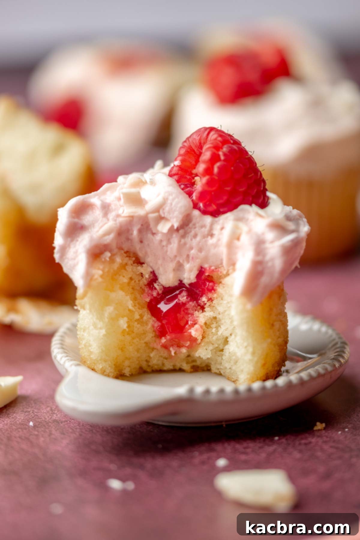 A beautifully decorated raspberry white chocolate cupcake with a bite taken out, revealing the raspberry filling inside.