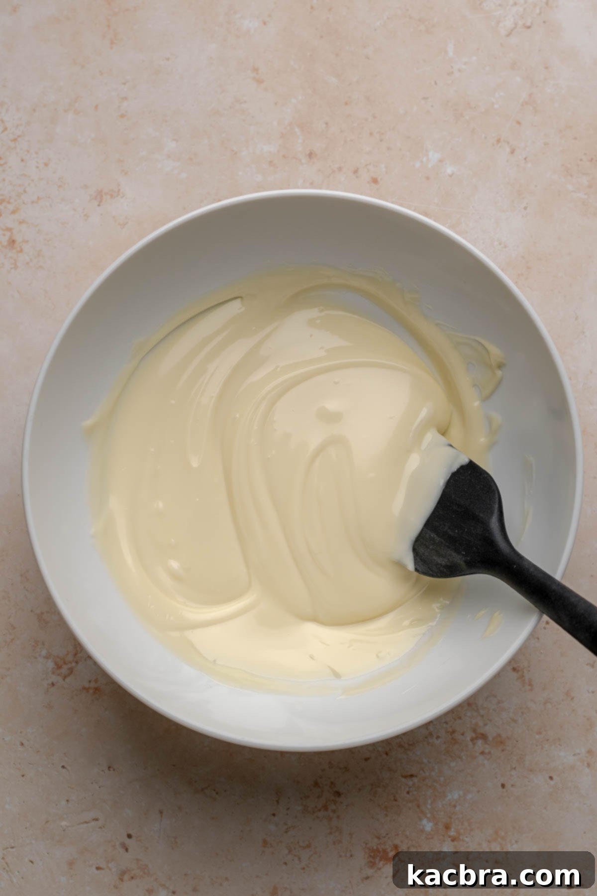 Smooth, melted white chocolate in a glass bowl with a spatula, indicating it's ready to be incorporated into frosting.