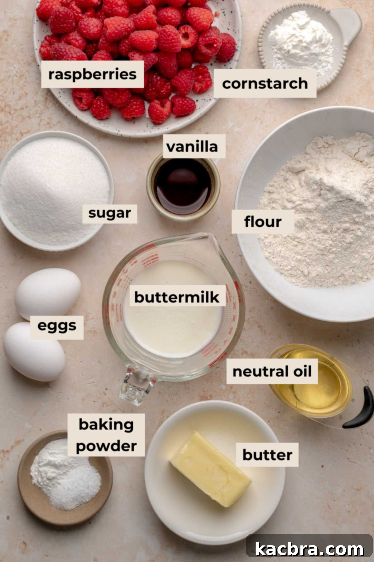 A flat lay photograph of various ingredients for cupcakes and raspberry filling, including flour, sugar, butter, eggs, buttermilk, and fresh raspberries.