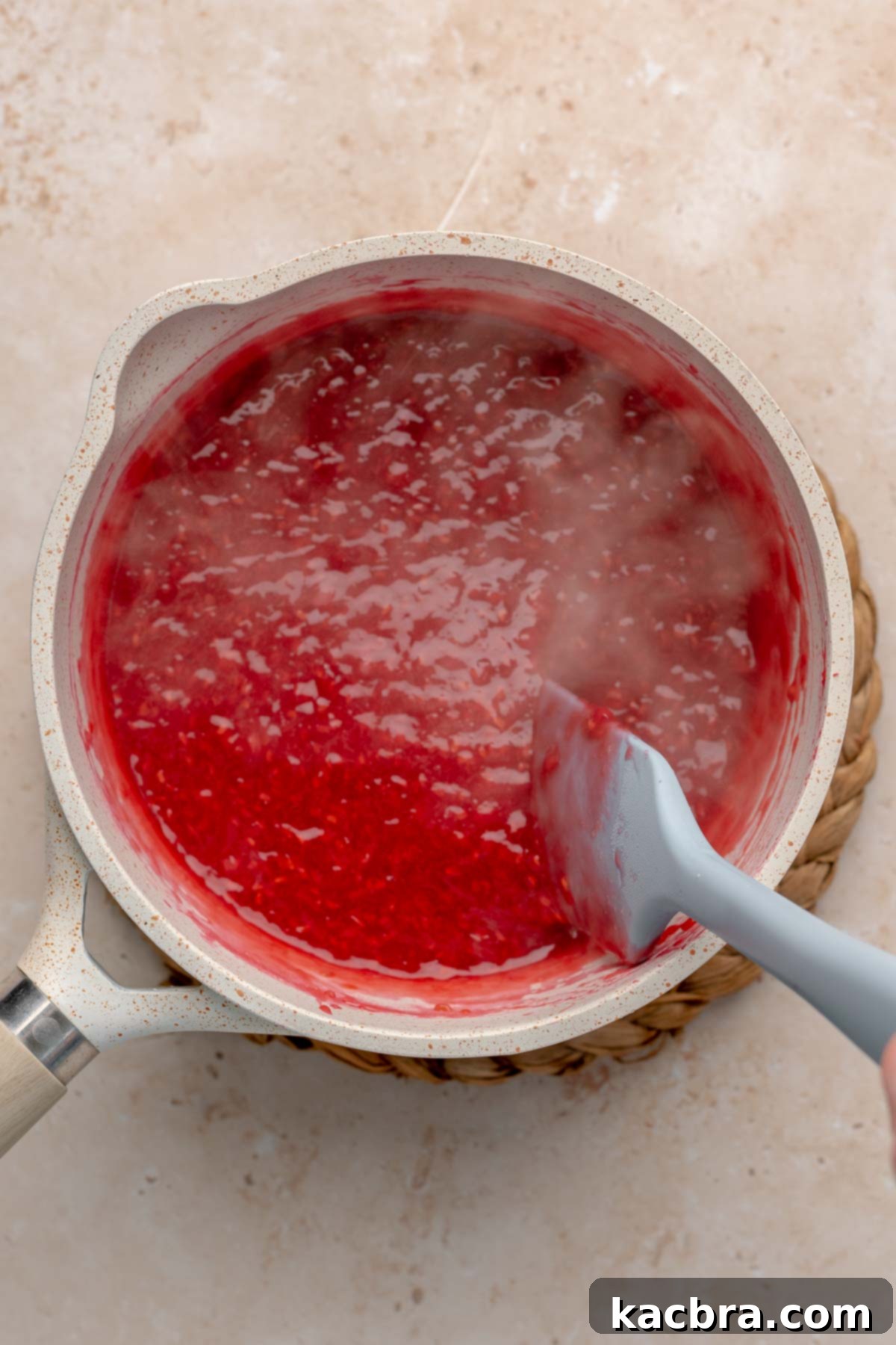 A spatula gently mixes hot raspberry puree in a saucepan on the stovetop, showing the fruit breaking down.
