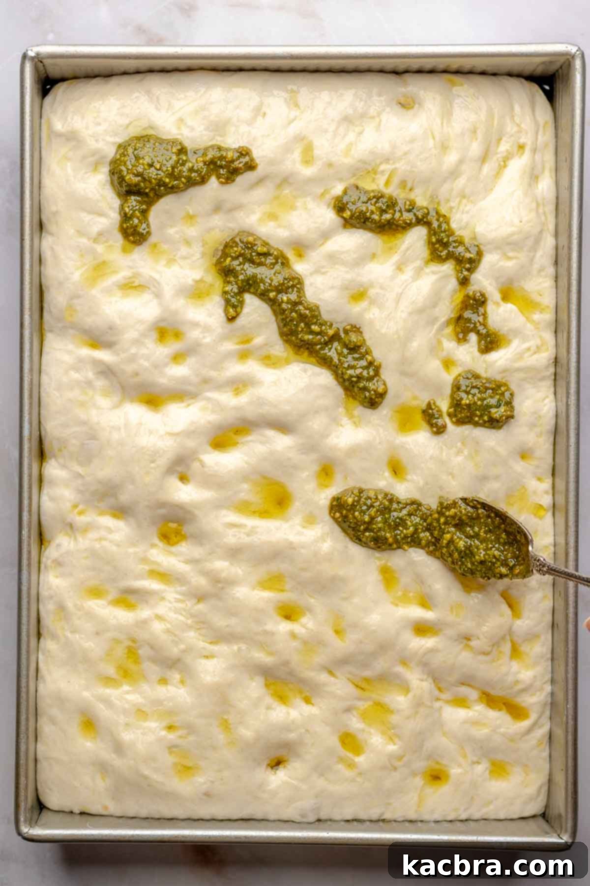 Aromatic Pesto Focaccia 14 A spoon adds pesto onto the risen focaccia dough in a pan.