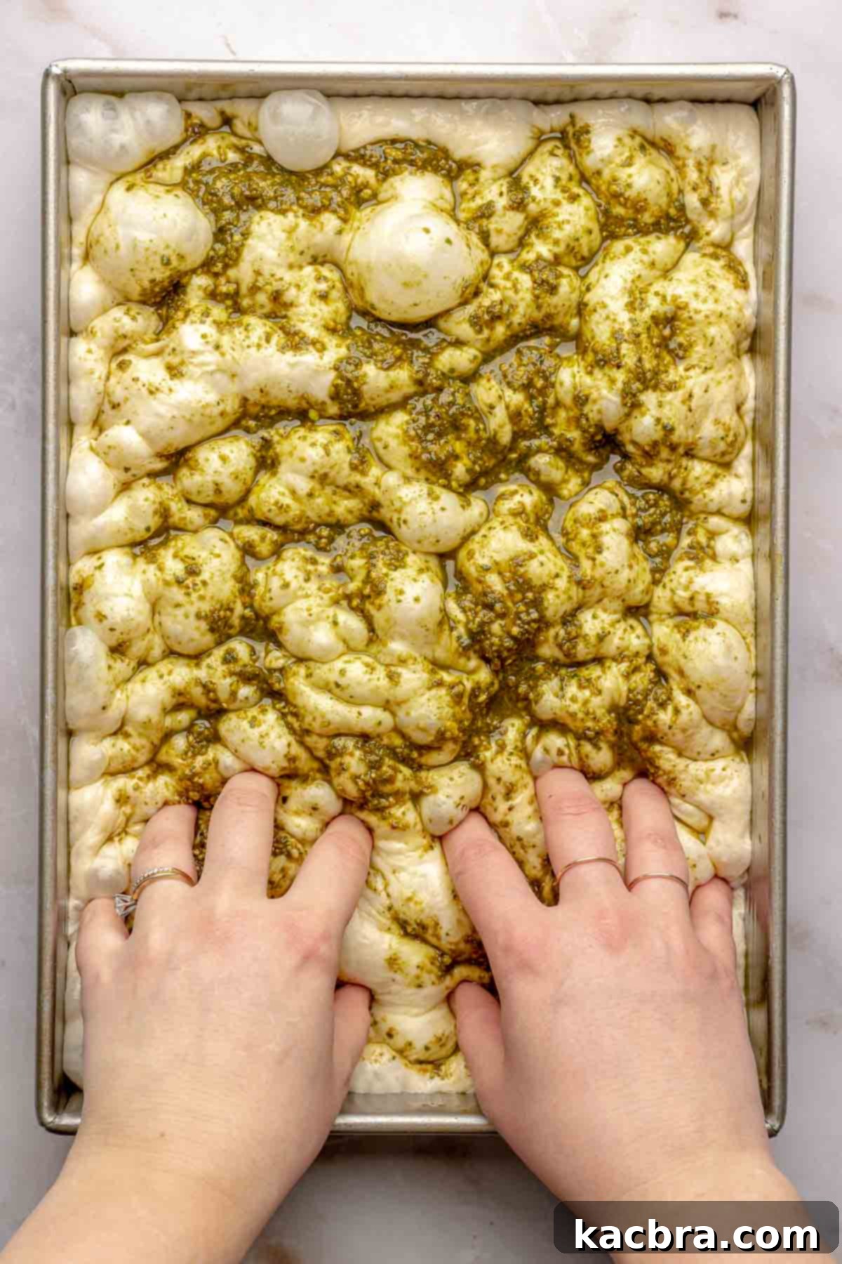 Aromatic Pesto Focaccia 15 Two hands press the pesto and dimples into the dough.