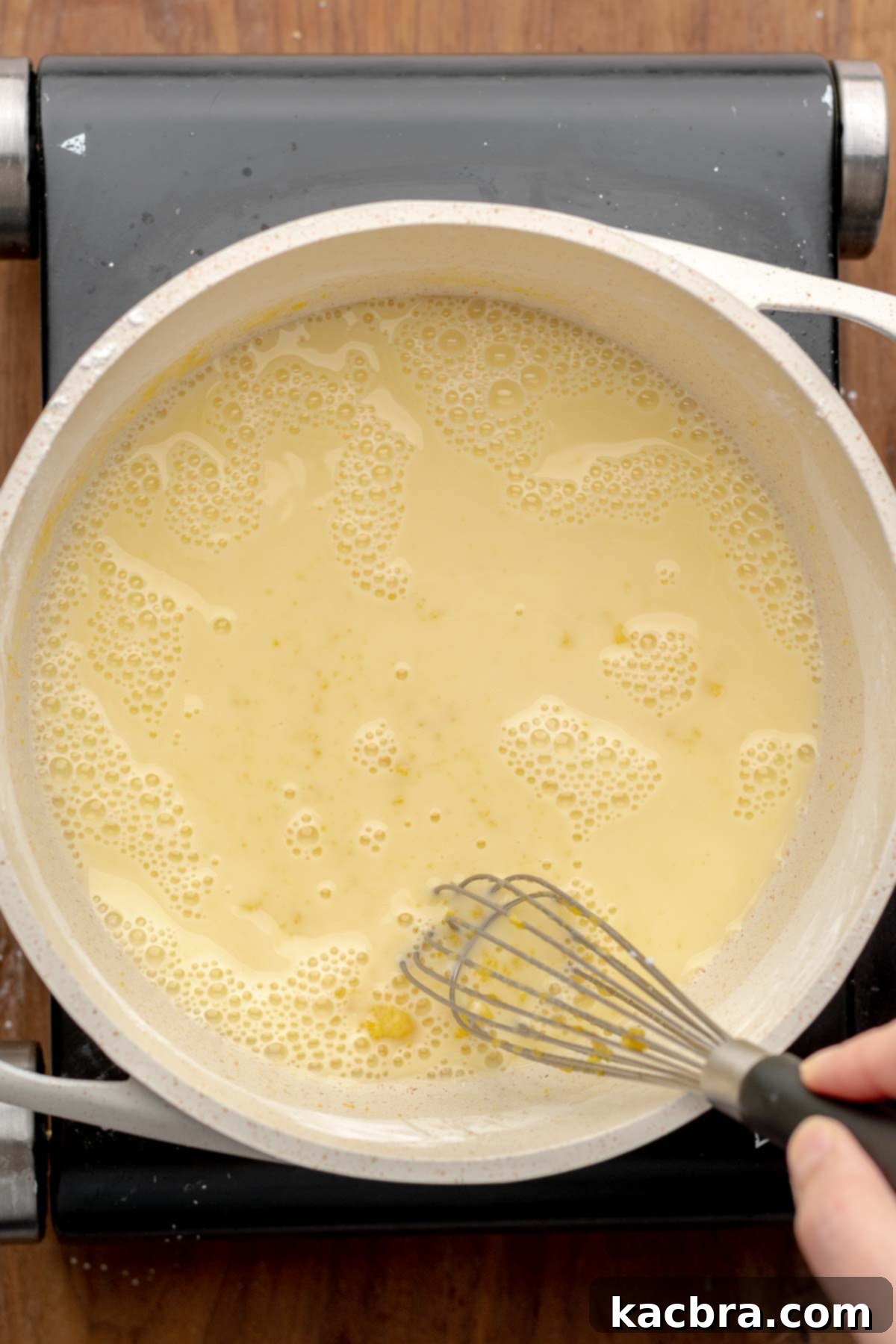 A hand whisks milk into the egg yolk and sugar mixture in a pot.