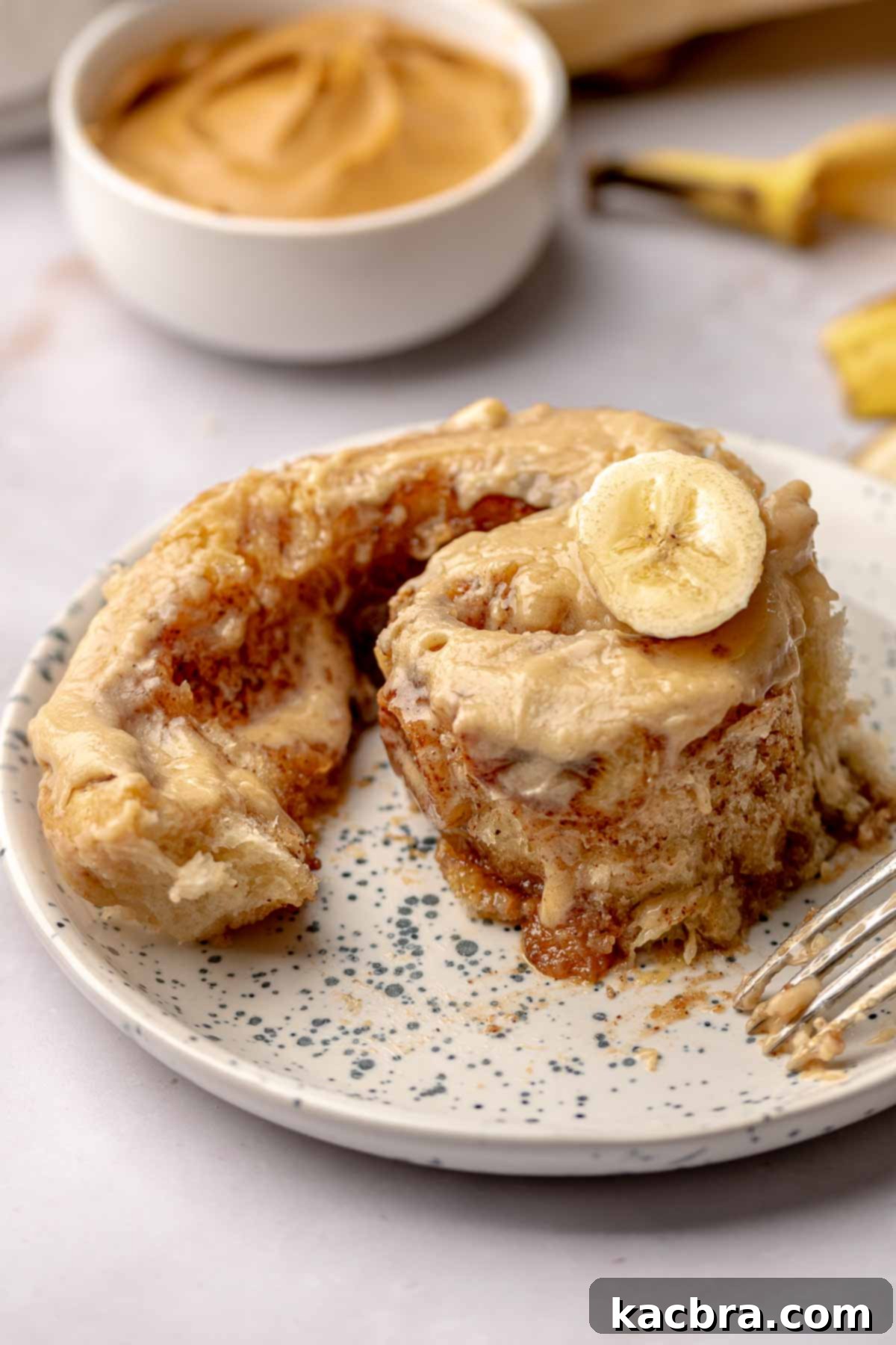 A perfectly unwound banana cinnamon roll, showcasing its gooey layers and peanut butter frosting, served on a white plate.