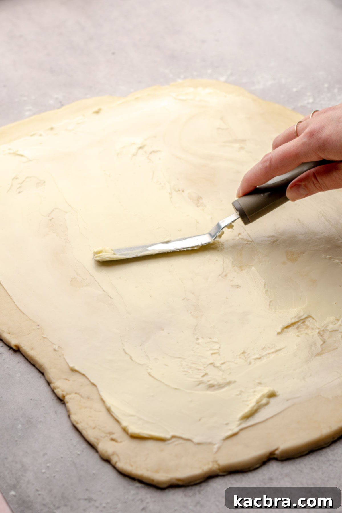A spatula spreads softened butter evenly across a sheet of rolled cinnamon roll dough.