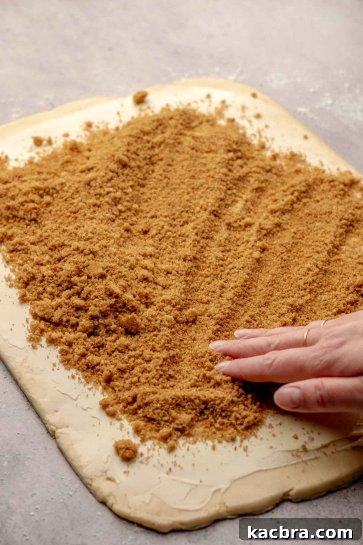 A hand sprinkles a mixture of cinnamon and brown sugar over the buttered dough.