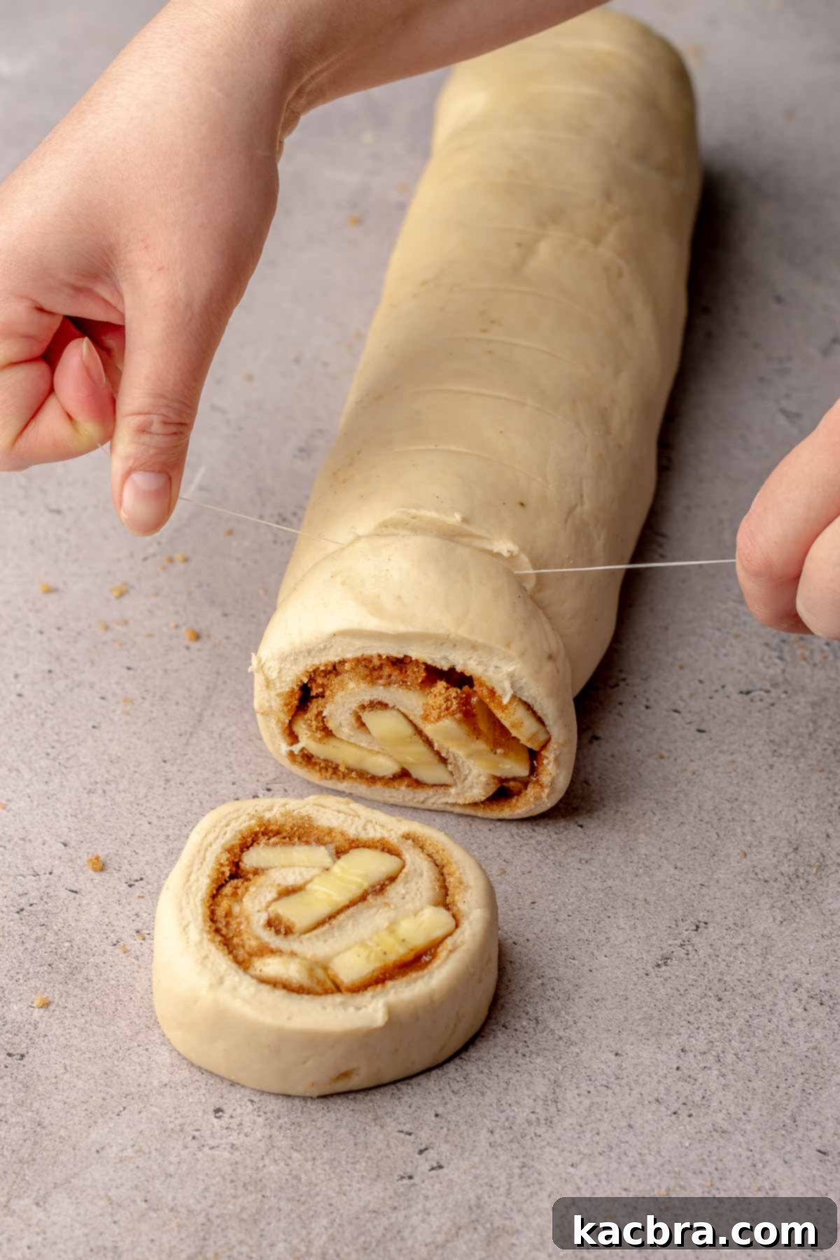 Hands demonstrating how to use dental floss to neatly cut a log of cinnamon rolls into individual portions.