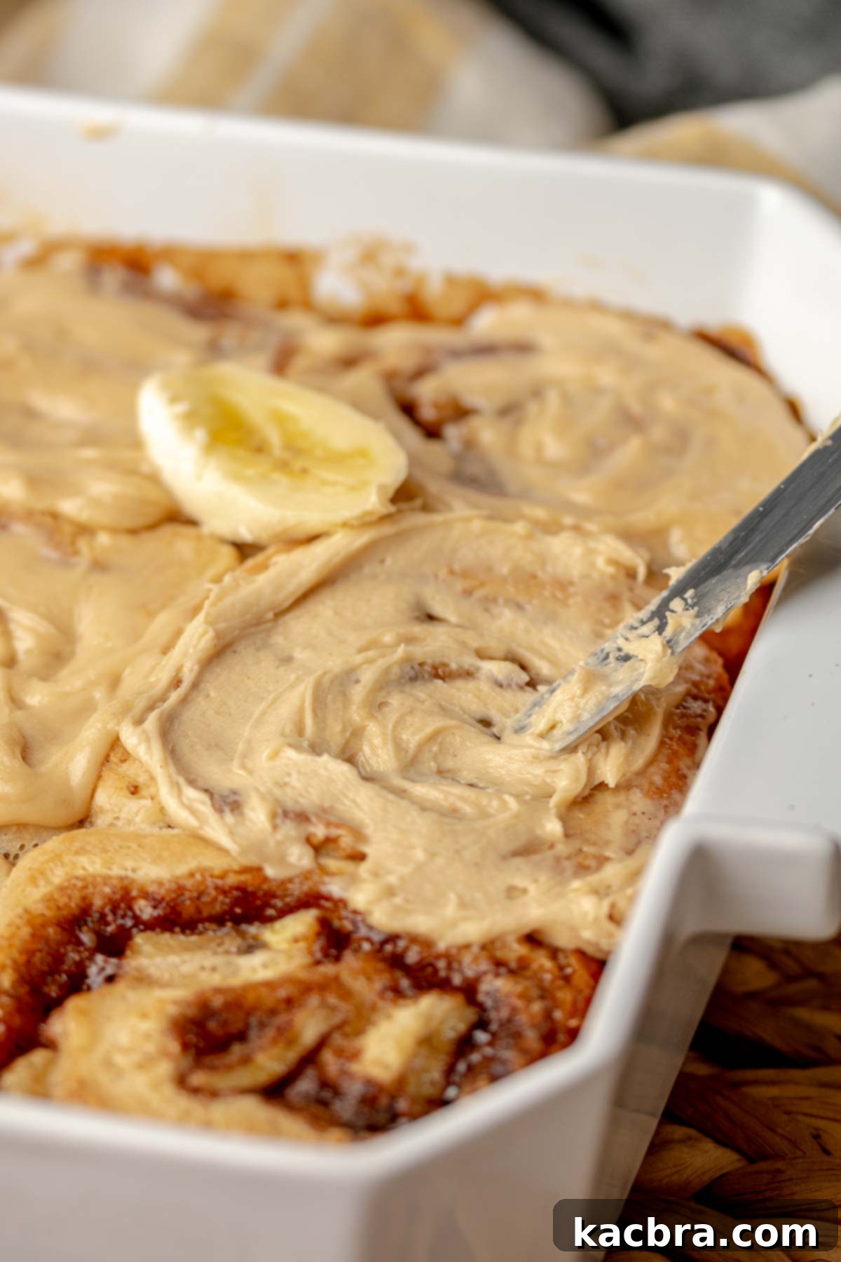 A spatula spreads luscious peanut butter frosting onto warm, freshly baked banana cinnamon rolls in their pan.