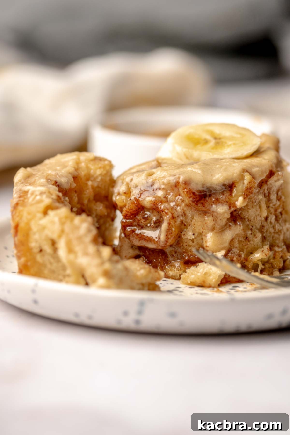 A half-unwound banana cinnamon roll on a plate, highlighting its soft, gooey interior and generous frosting.
