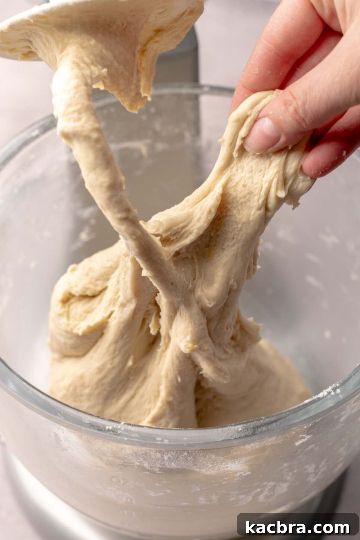 A hand gently pulls finished cinnamon roll dough off of the dough hook, showing its smooth, elastic texture.