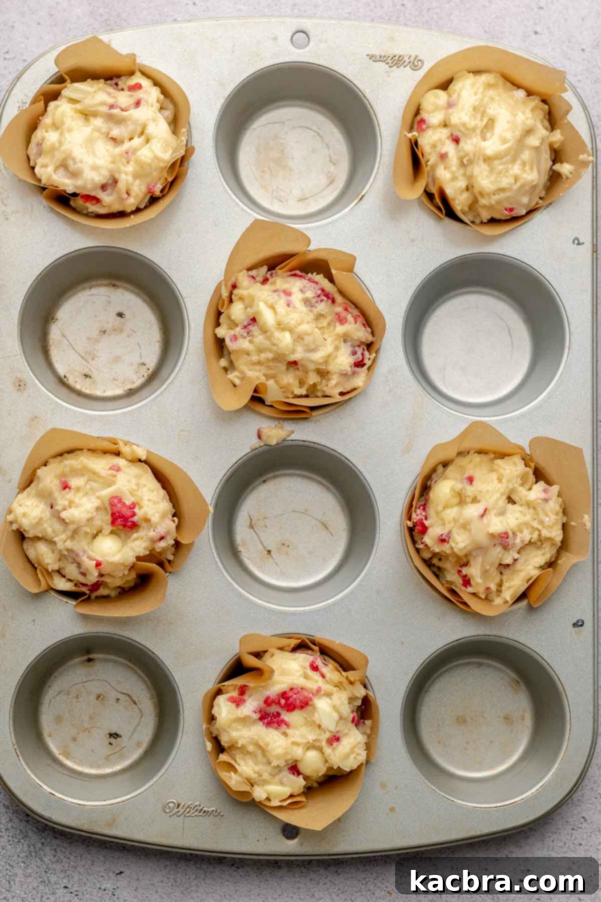 A muffin tin with six liners filled to the brim with white chocolate raspberry muffin batter, strategically placed for optimal baking.