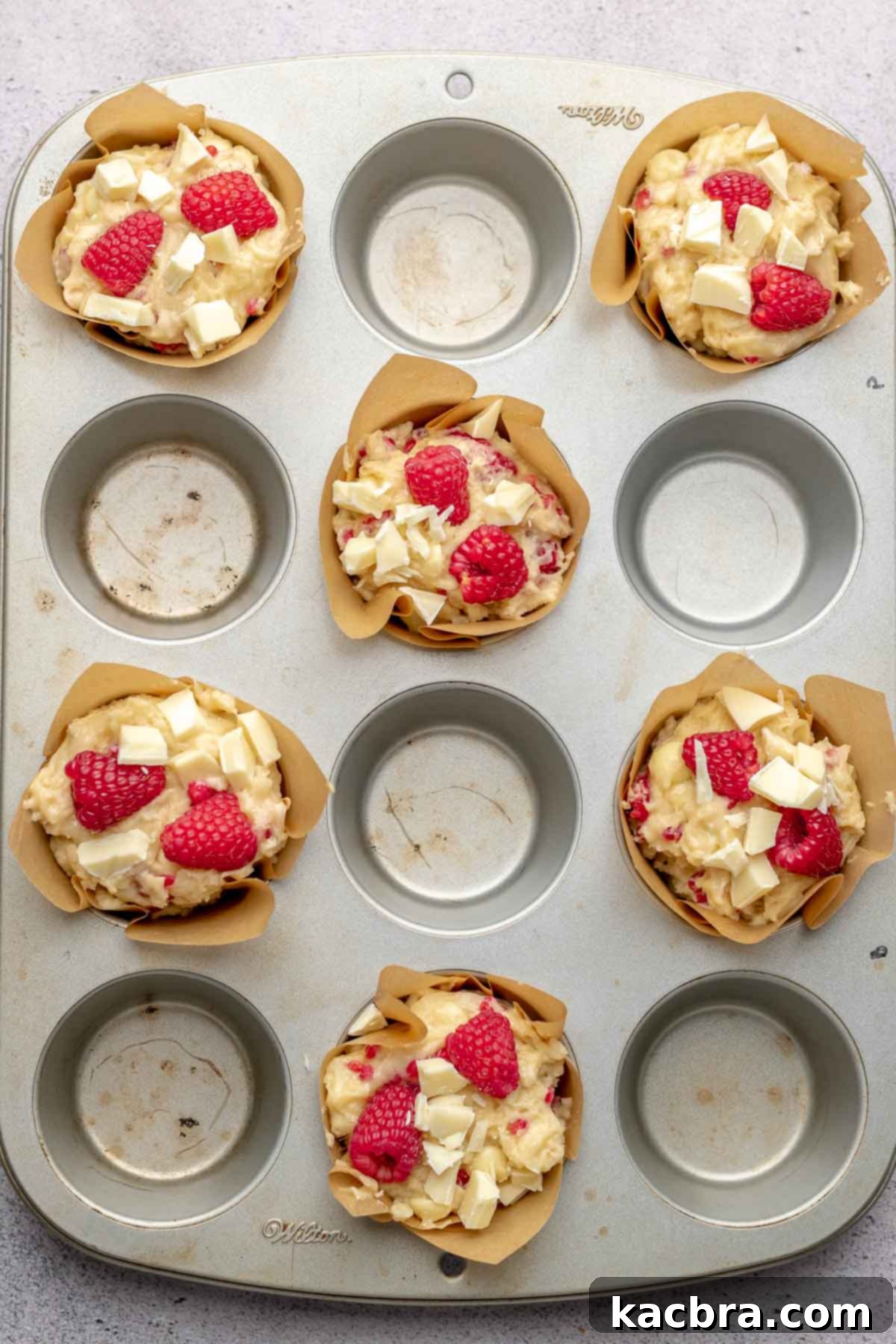 Unbaked muffins generously topped with extra fresh raspberries and white chocolate pieces, promising delicious bursts of flavor and a beautiful presentation.